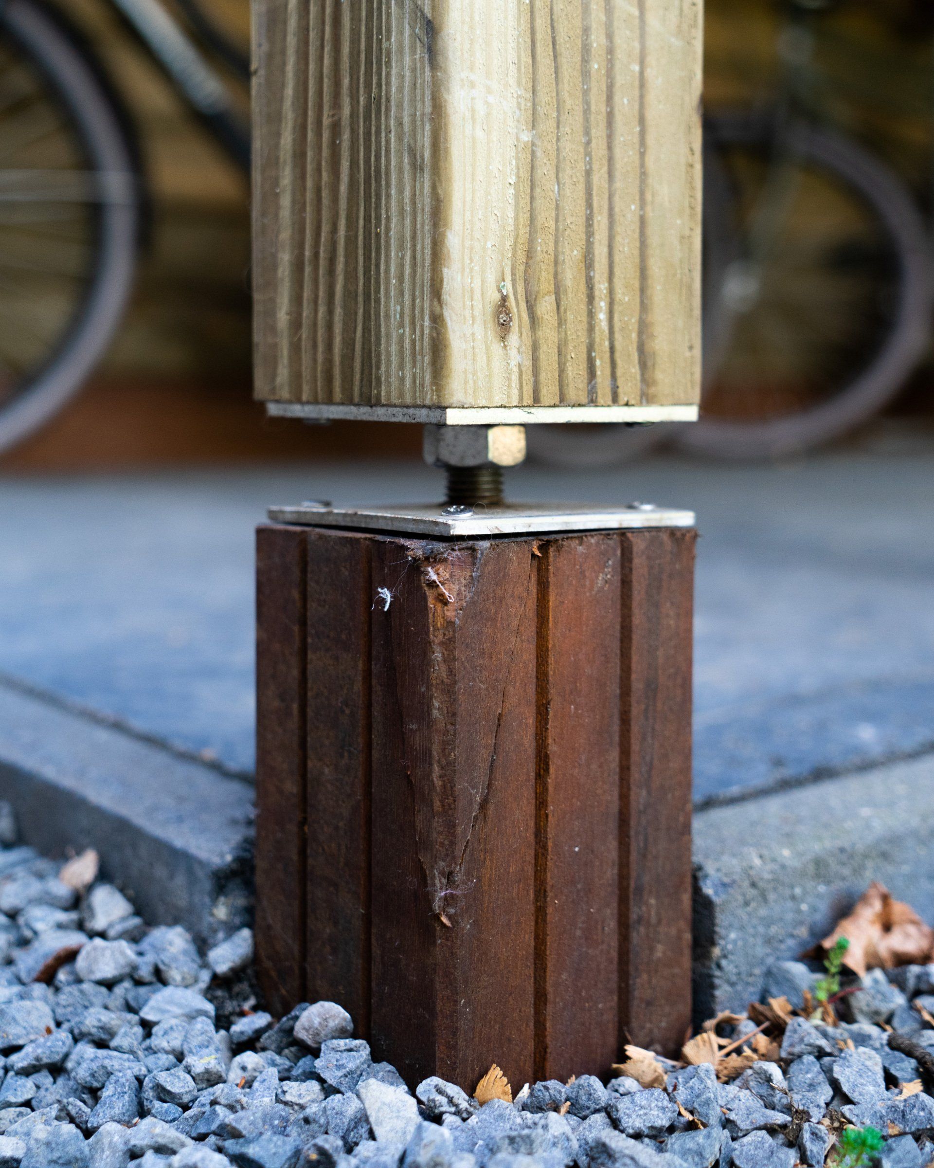 Houten paalconstructie: een vierkante, lichtgekleurde paal bovenop een donkere, cilindrische voet, verbonden door metalen bevestigingsmaterialen.