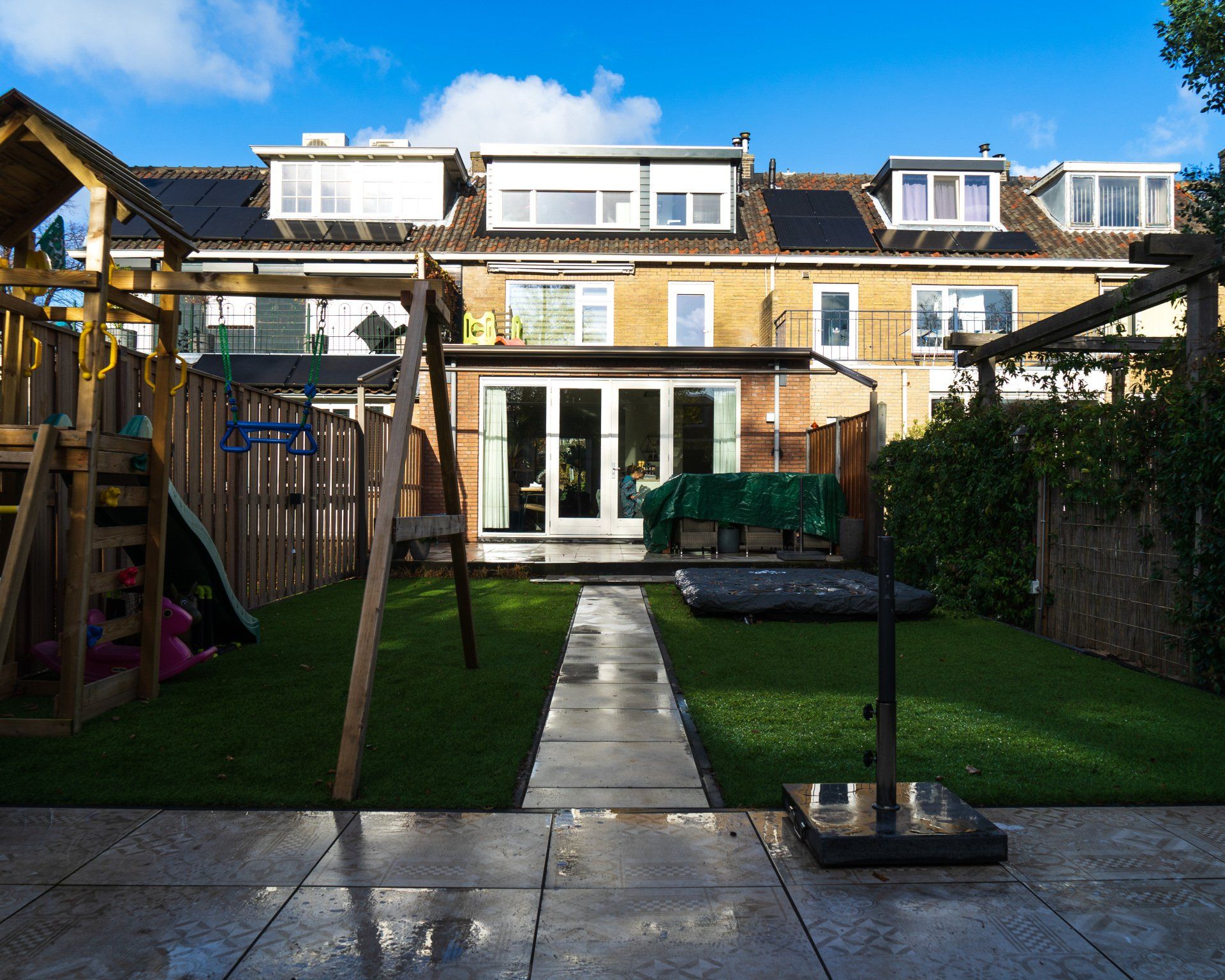 Achtertuin met groen gazon, speeltoestel, terras en een rij huizen.