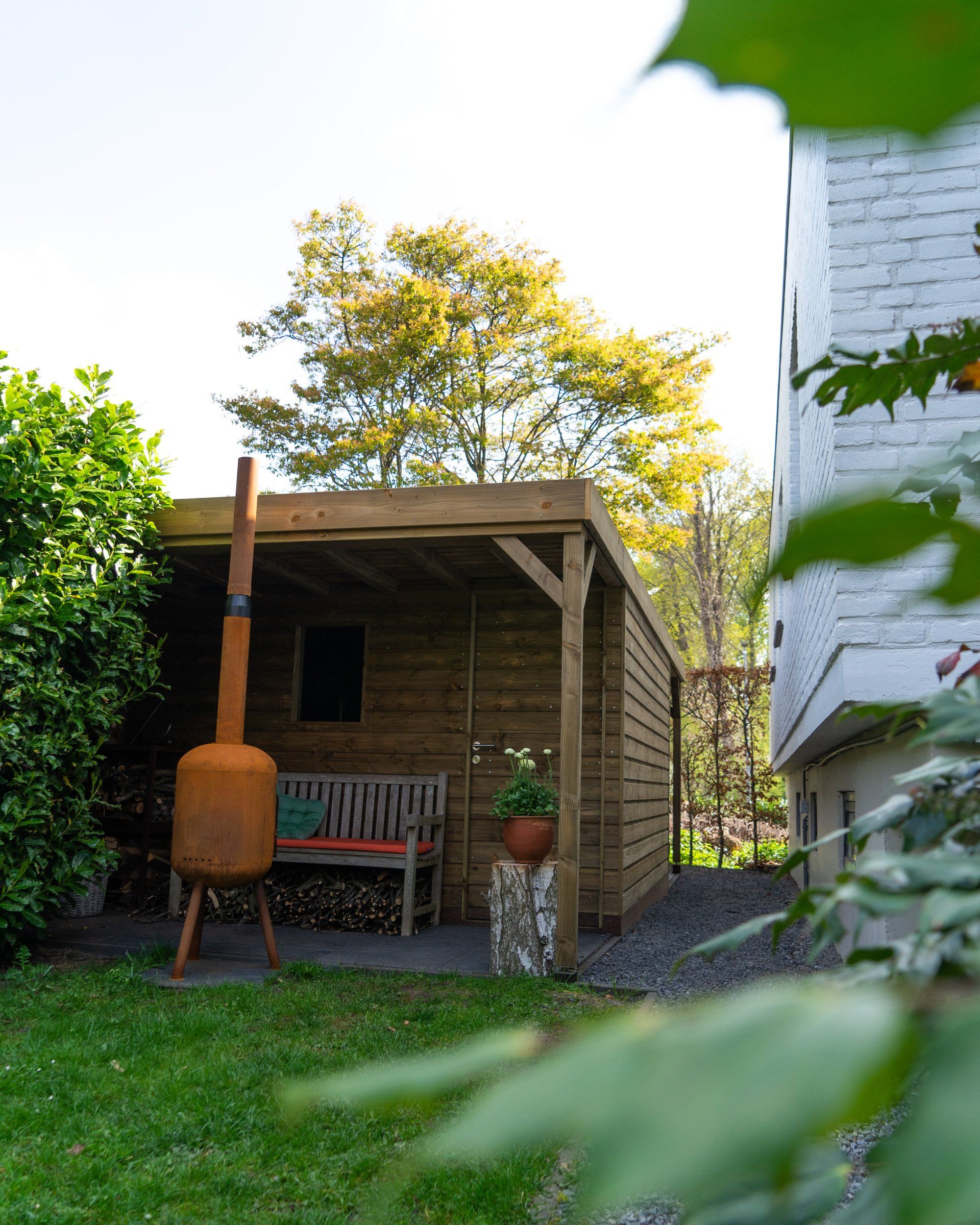 Houten schuur met bankje, roestige buitenhaard en groen.
