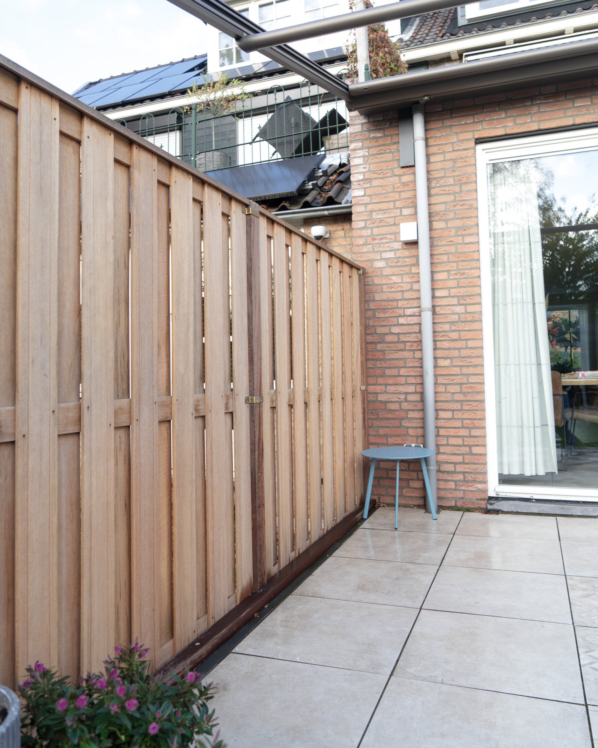 Een houten schutting naast een bakstenen muur met een glazen schuifdeur en een klein stoeltje op een terras.