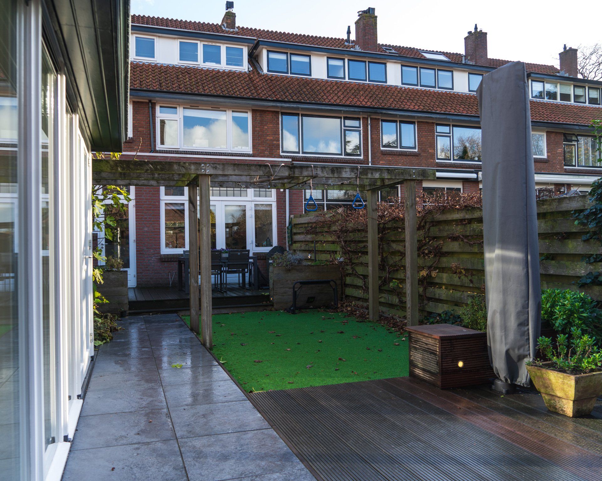Terras in de achtertuin met pergola, kunstgras en een aangrenzend gebouw met rood dak.