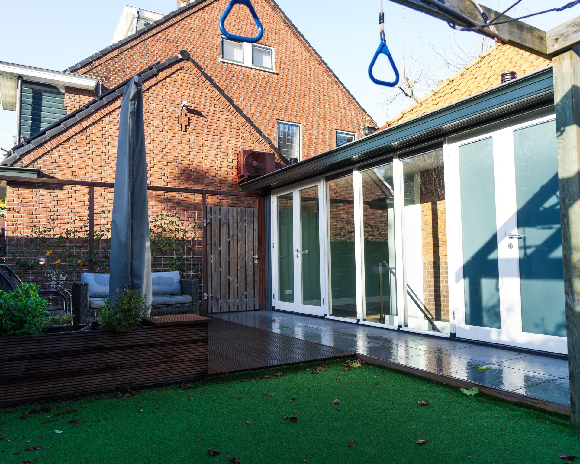 Achtertuin met bakstenen huis, glazen deuren en schommel. Groen gazon, houten terras en grijze pilaar.