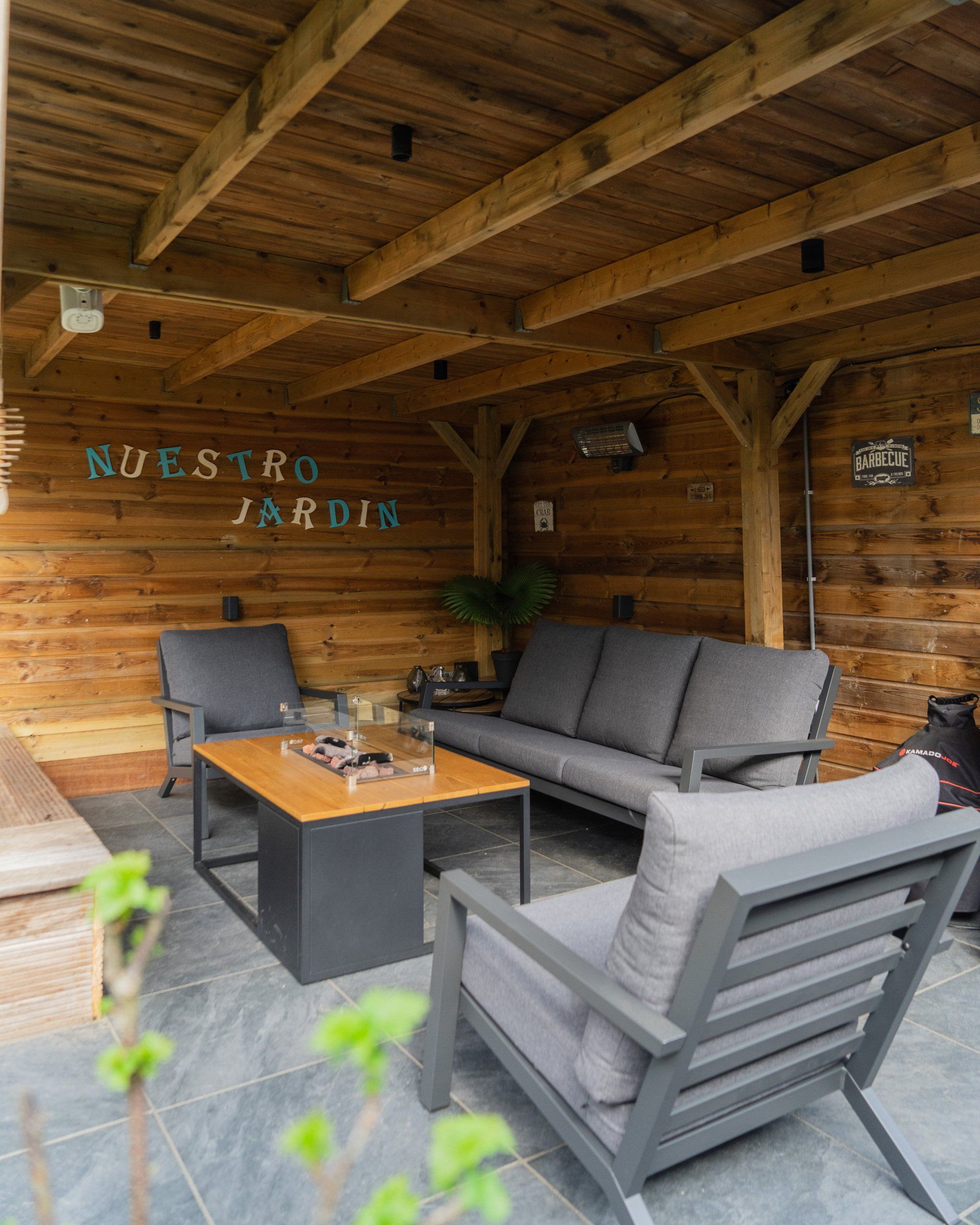 Buitenpatio met houten balken, grijze meubels, een salontafel en de woorden 
