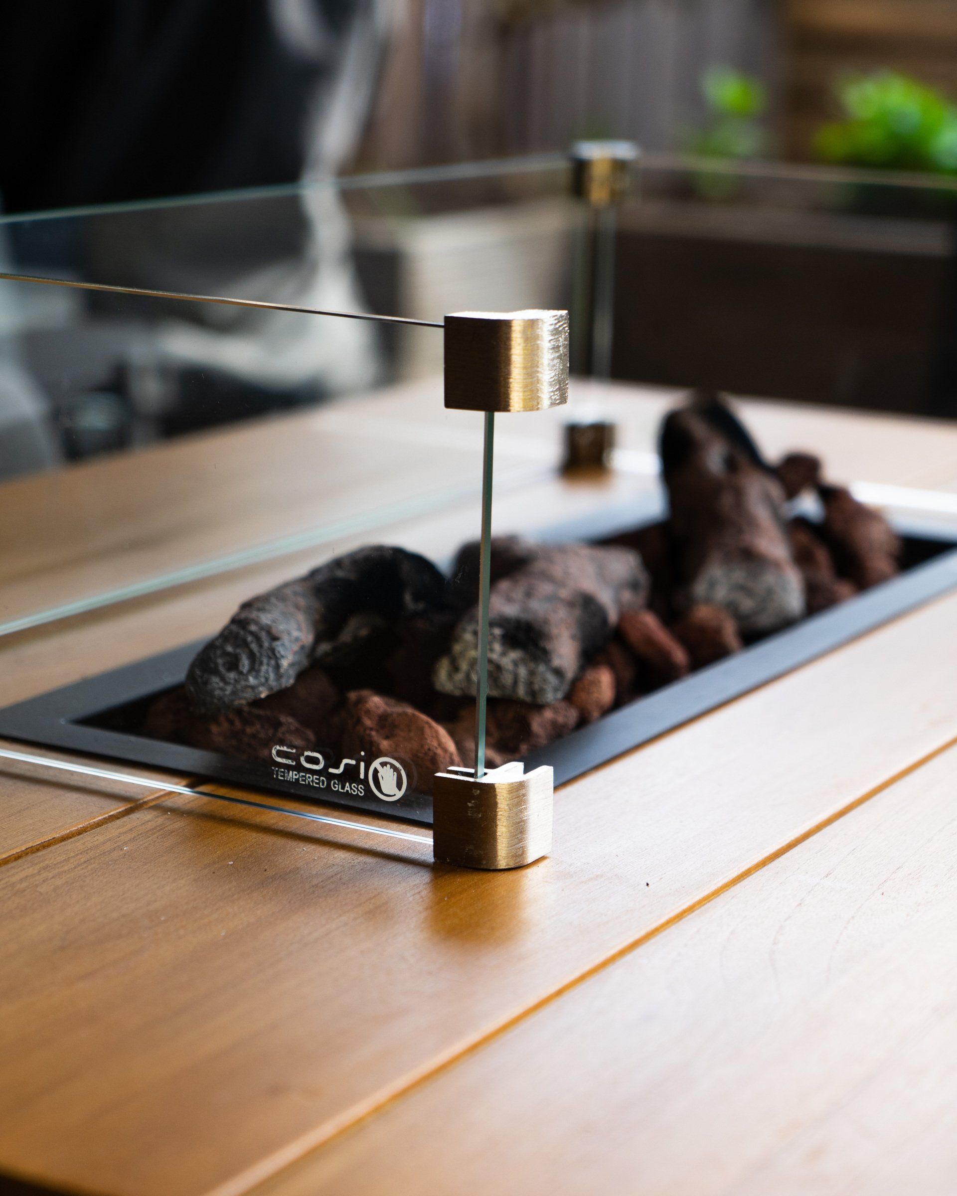 Detailopname van een moderne buitenhaard met glazen windscherm, houten tafel en lavastenen.