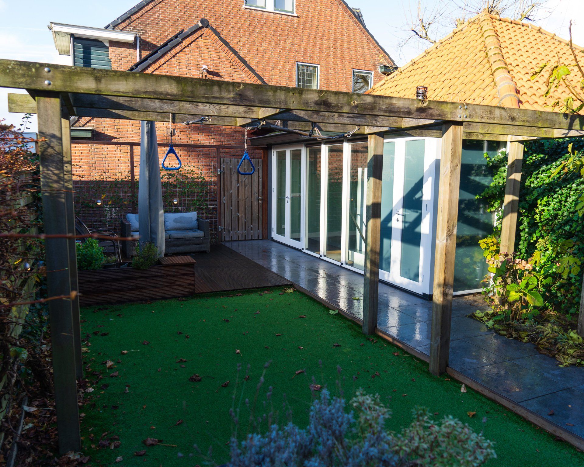 Terras in de achtertuin met houten pergola, kunstgras en glazen deuren, grenzend aan een huis.