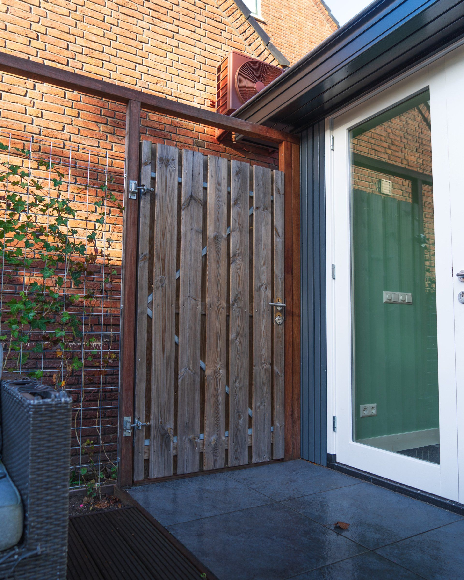 Houten poort in een frame van baksteen en metaal, die toegang geeft tot een glazen deur met groene wanden.