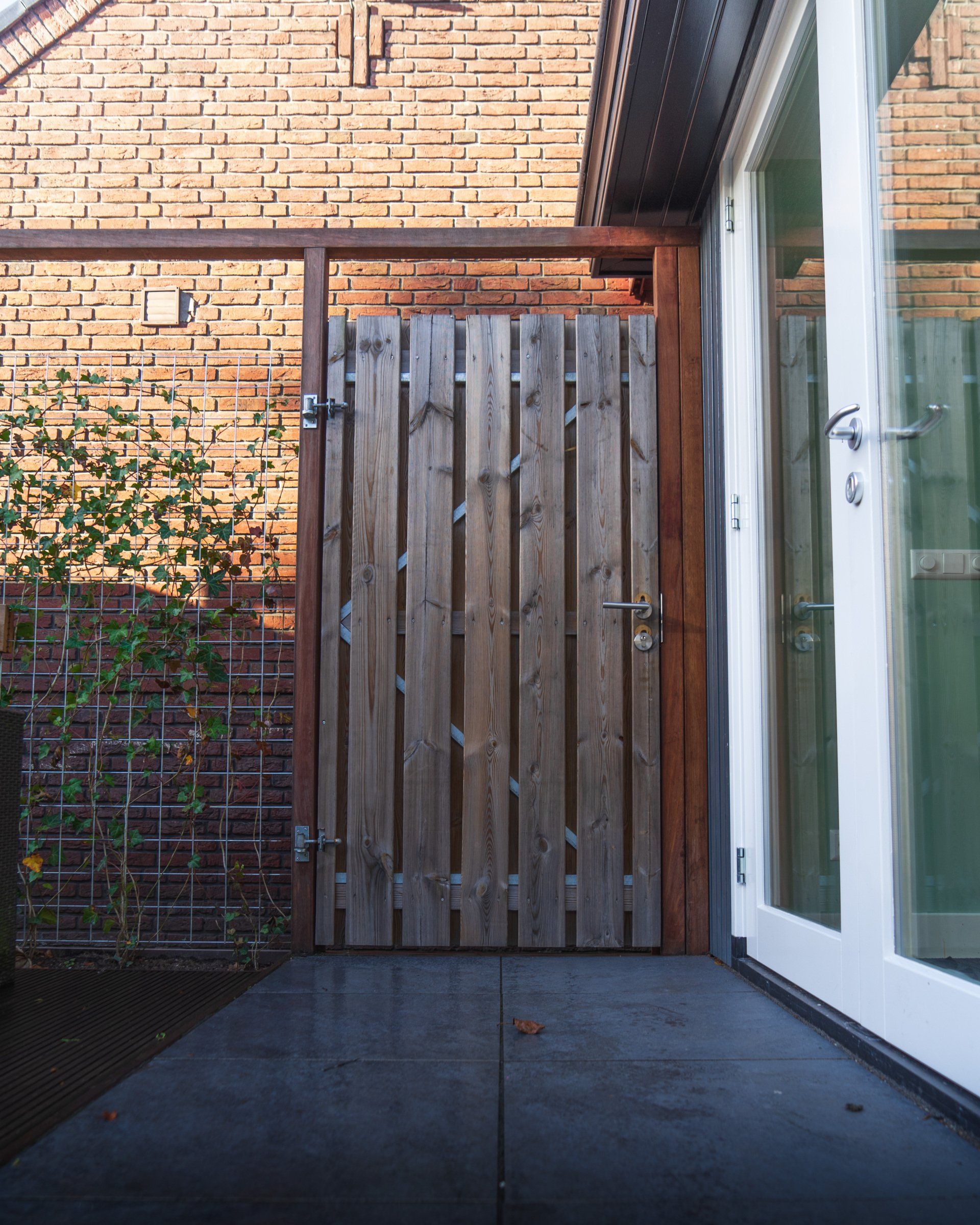 Houten poort in een binnenplaats met bakstenen muren. Rechts daarvan een glazen deur.