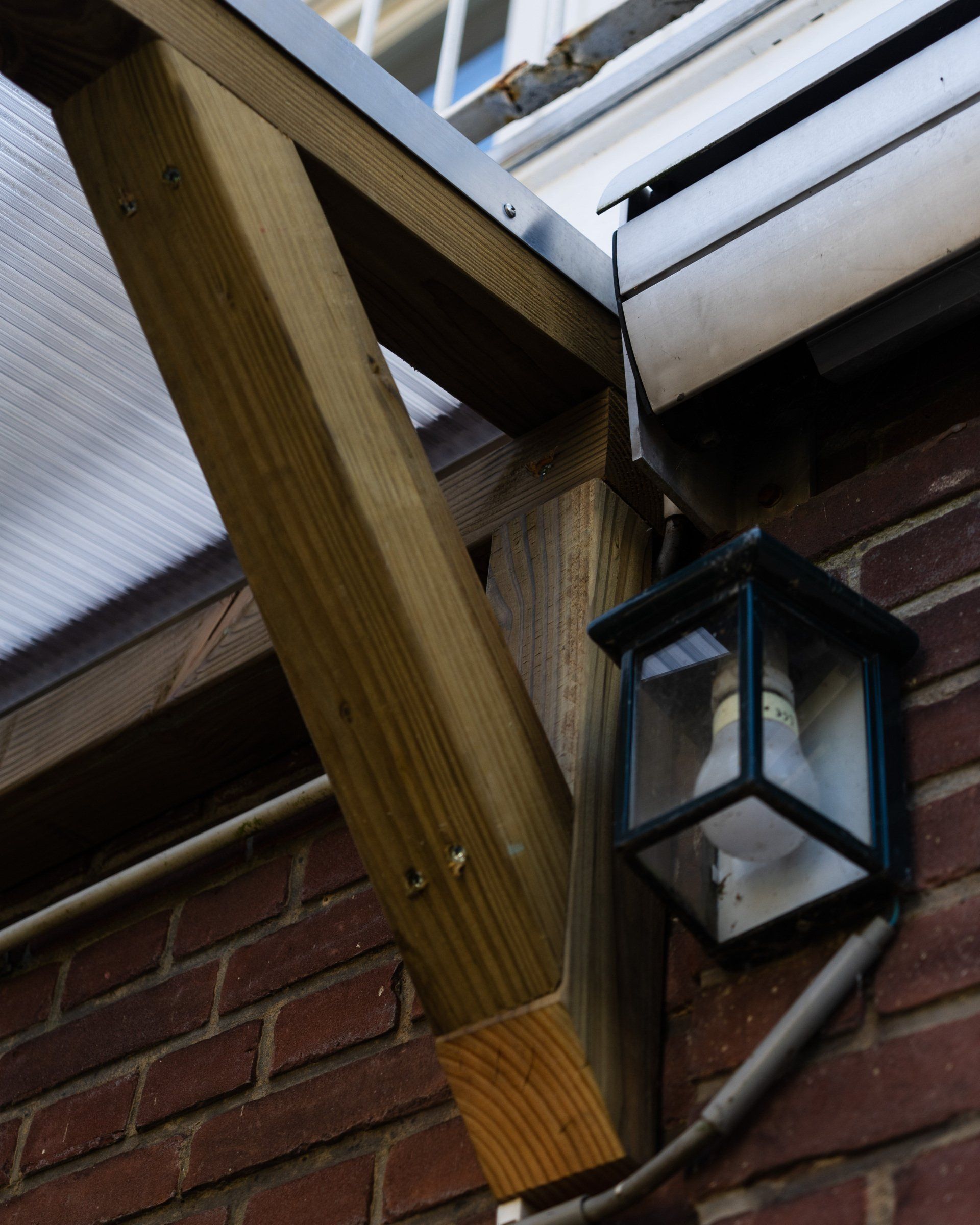 Houten draagconstructie bevestigd aan een bakstenen muur, met een kleine buitenlamp en transparante dakpanelen.