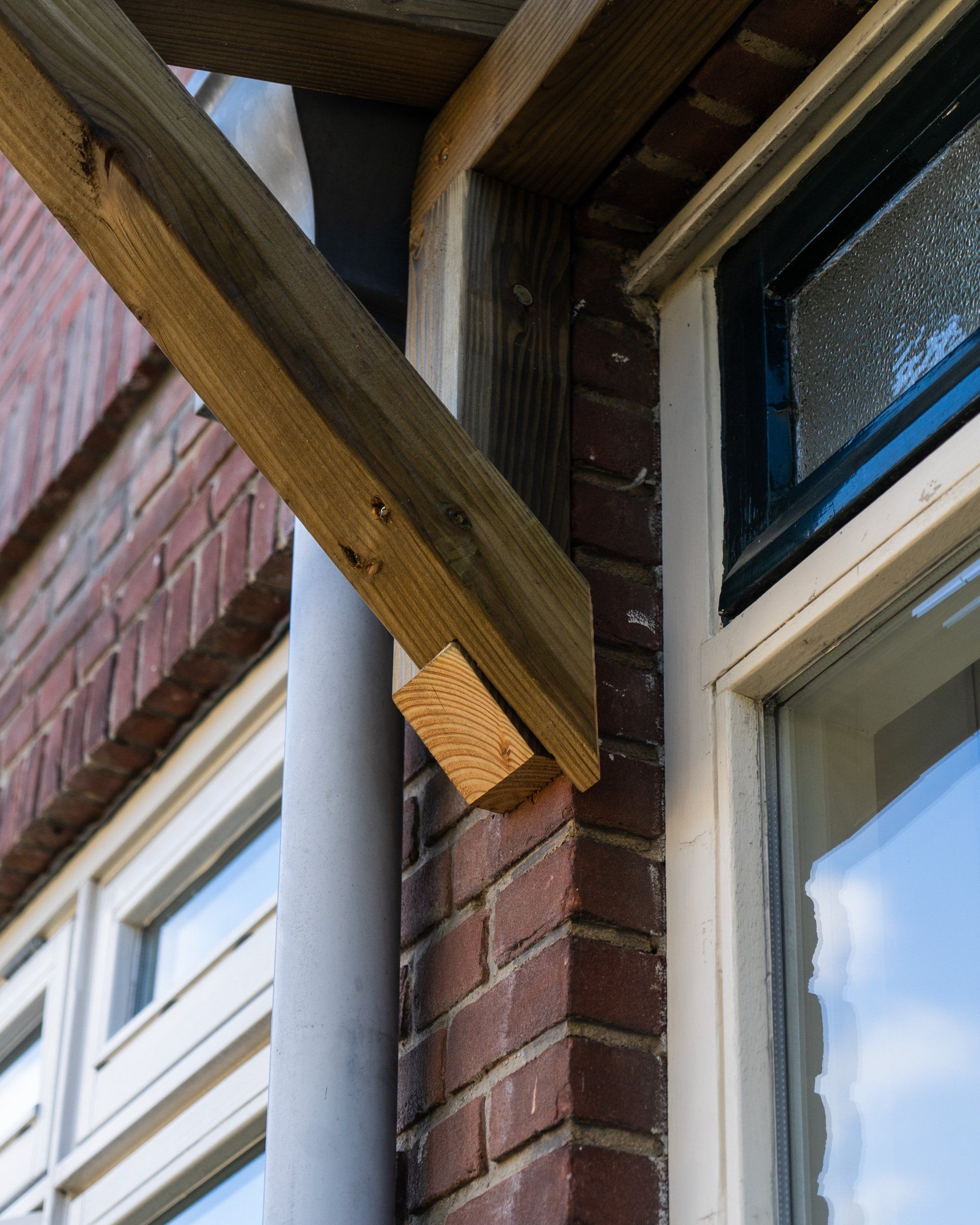 Houten balk die een dakoverhang ondersteunt, bevestigd aan een bakstenen gebouw naast een raam.