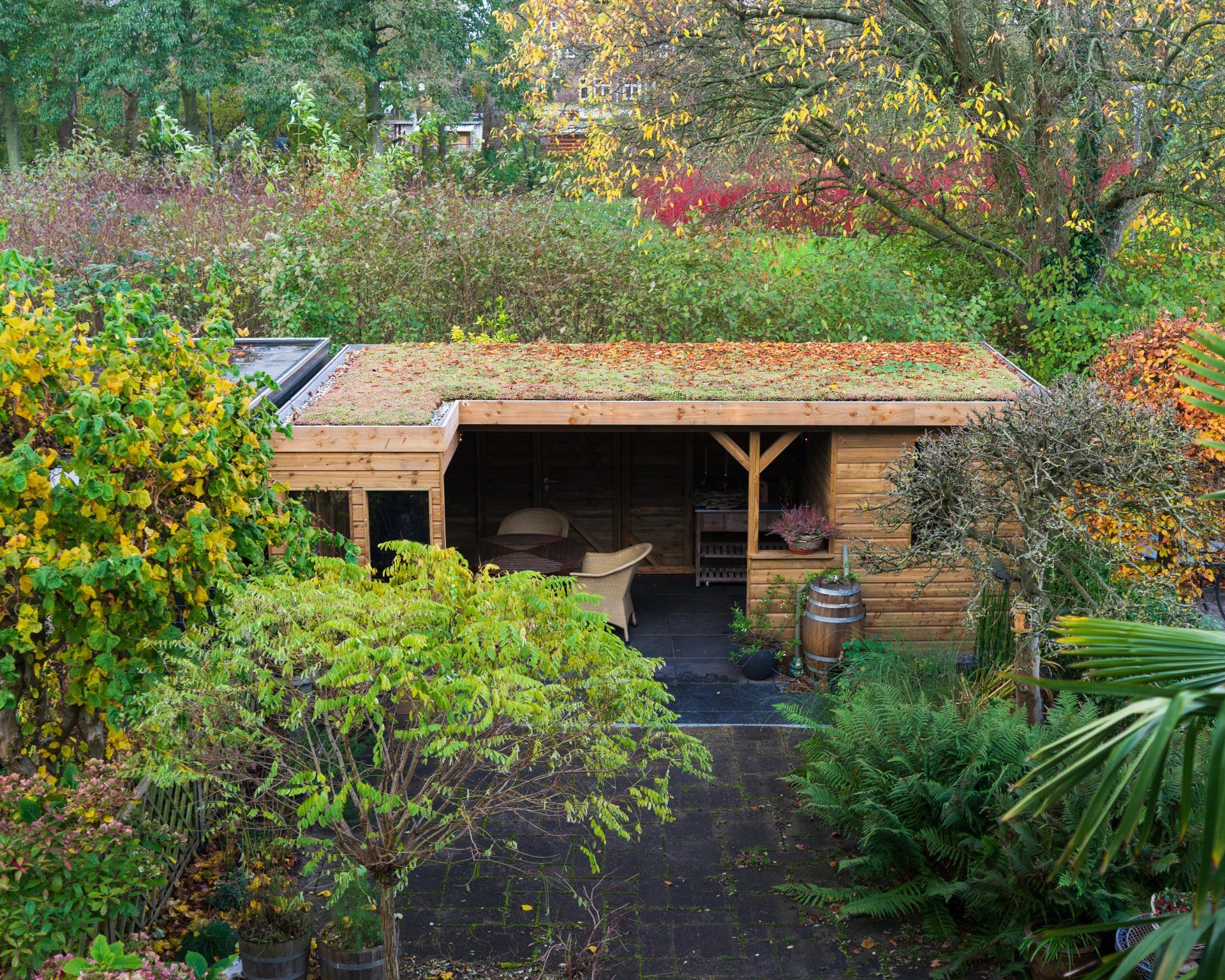 Houten schuur met een groen dak, in een weelderige tuin. Bomen omringen de open constructie.