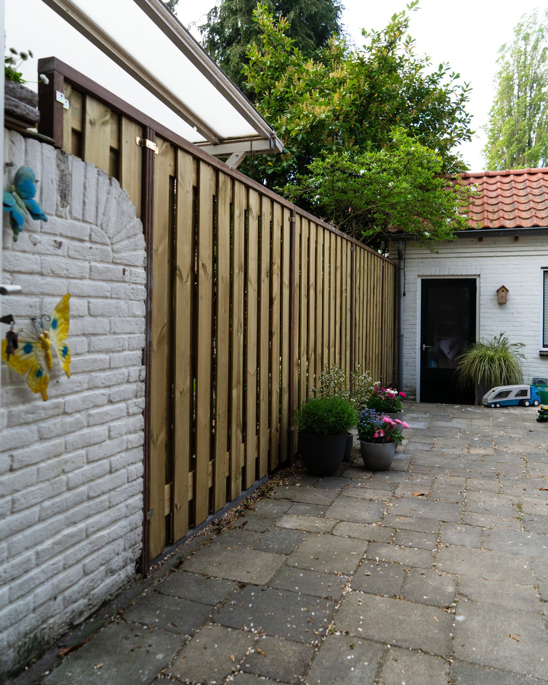 Een houten schutting naast een bakstenen muur leidt naar een terras met planten en een deur.