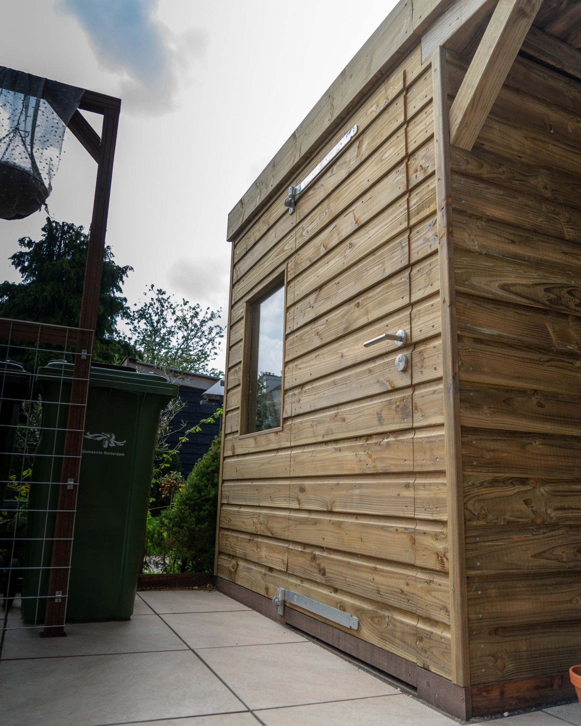 Houten schuur met horizontale zijwanden, een klein raam en een open deurklink, gelegen op een terras.