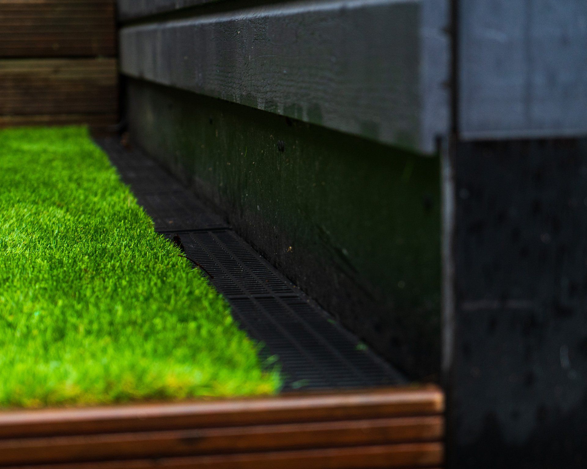 Close-up van een laagliggend groen kunstgrasveld naast een donkerhouten hek en een afvoerrooster.