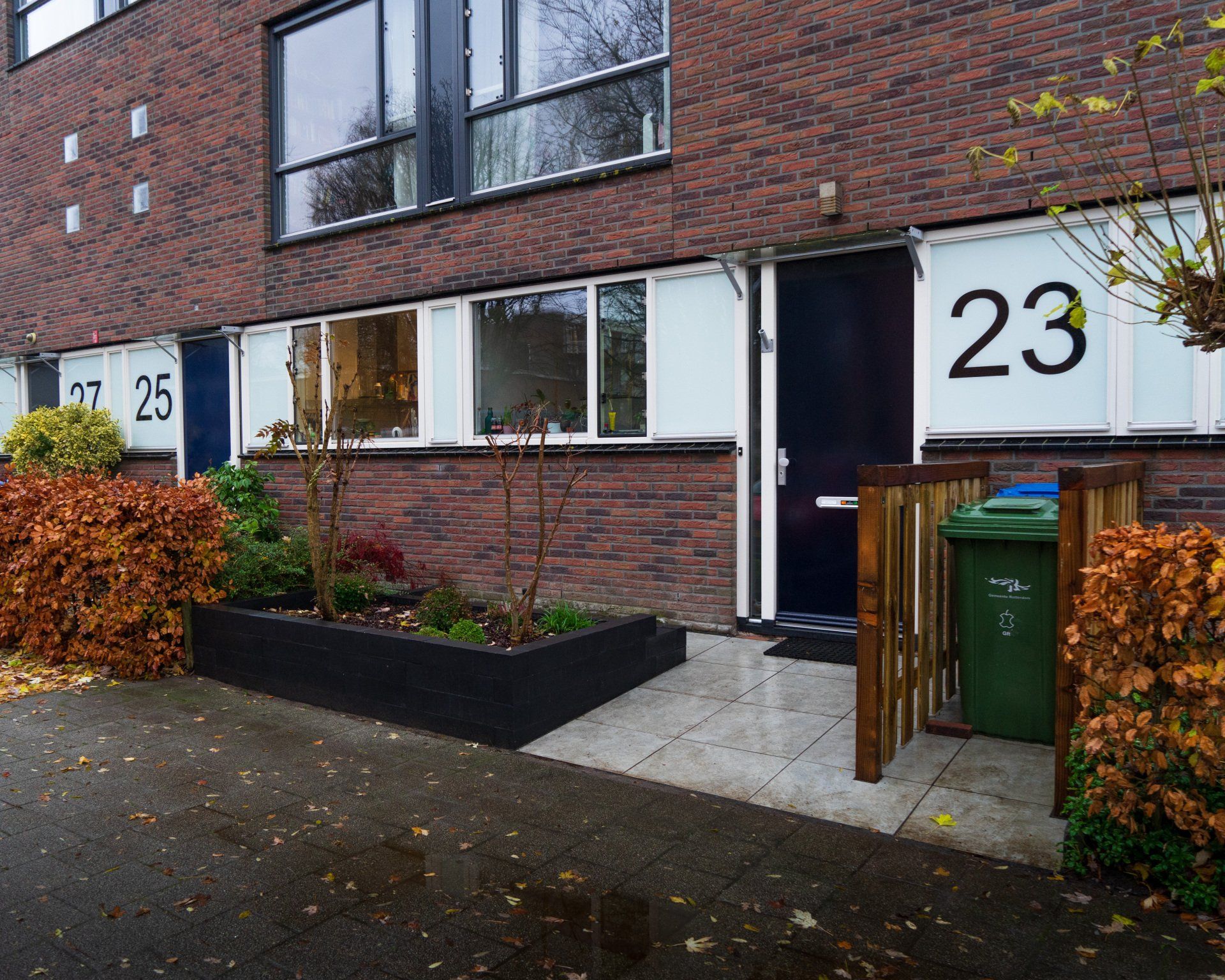 Rij bakstenen rijtjeshuizen, huisnummers 23 en 25 zichtbaar. Vooringangen met tuinen en een vuilnisbak.