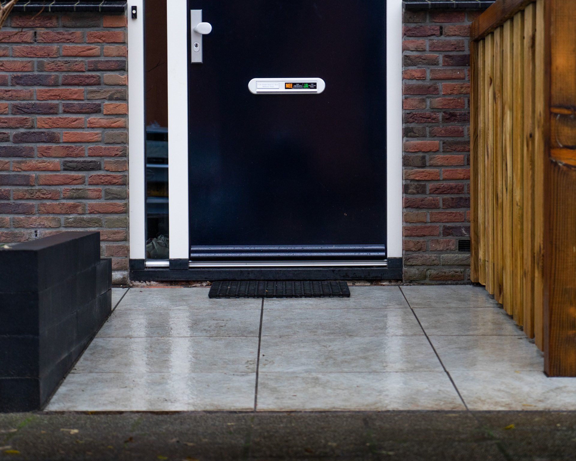 Zwarte voordeur met witte kozijnen, bakstenen muur, betonnen pad en houten schutting.