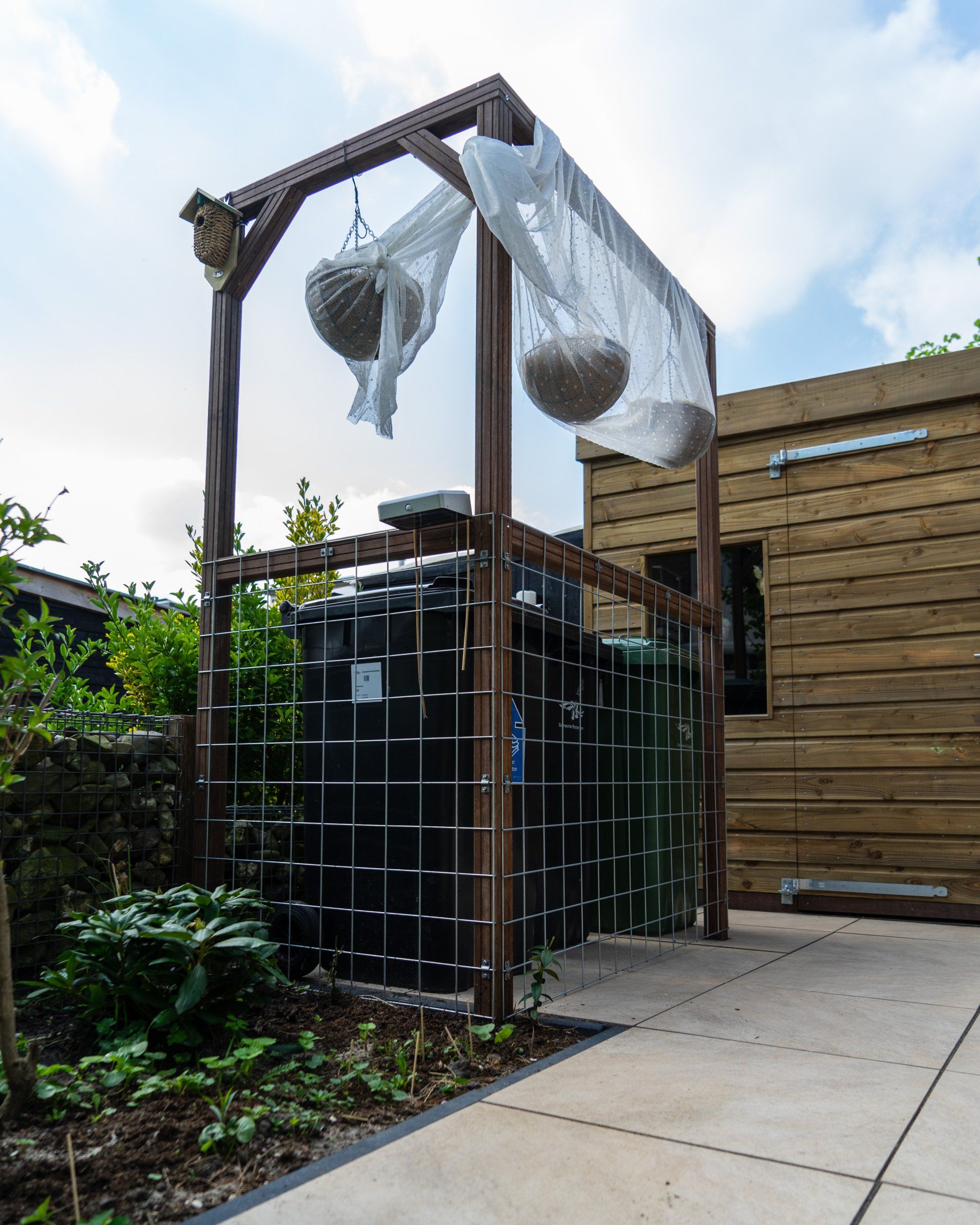 Een houten frame beschermt een zwarte watertank. Twee zaadbollen hangen aan de bovenkant. Op de achtergrond staat een houten schuur.