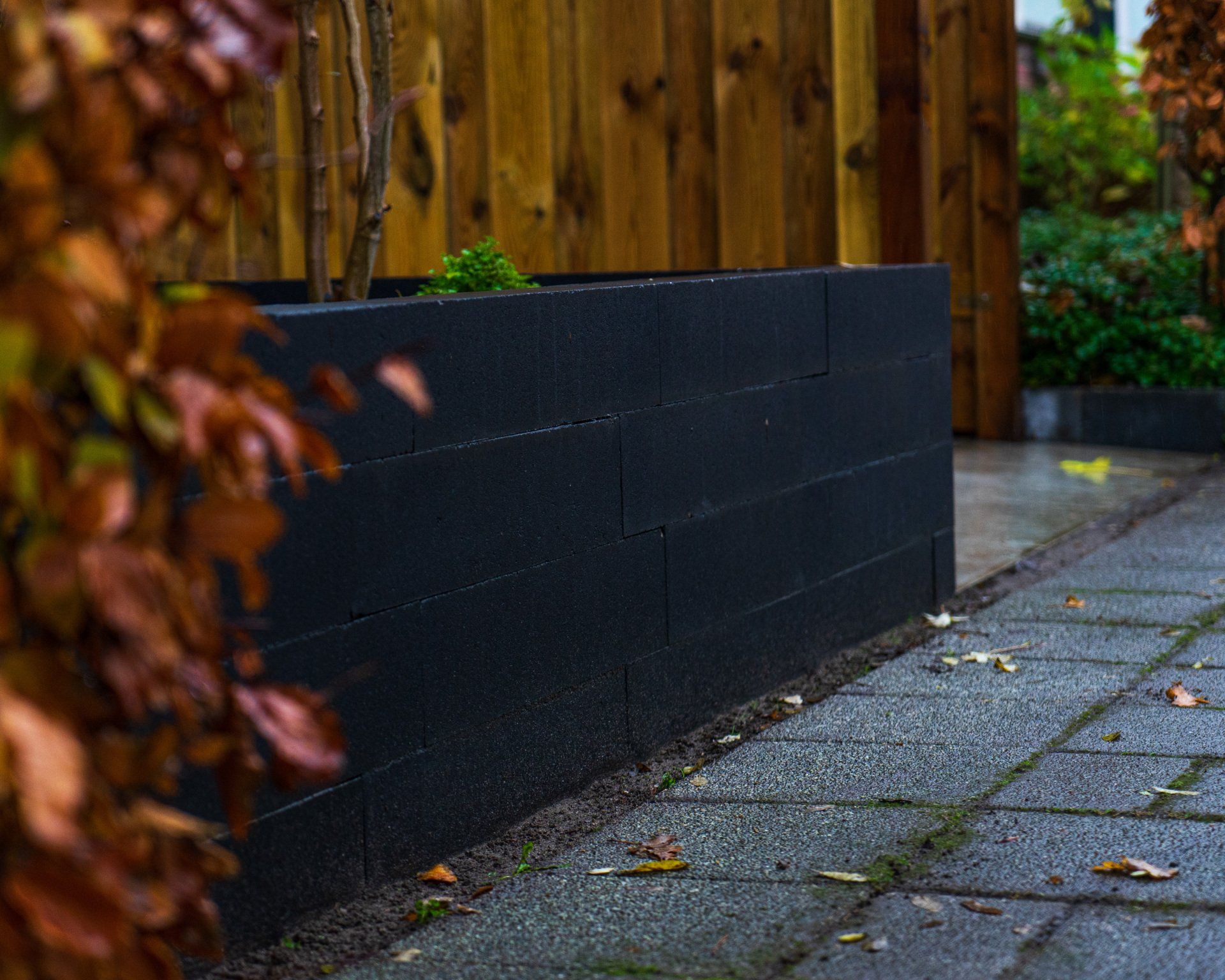 Zwarte, rechthoekige plantenbak langs een bakstenen pad, met een houten schutting op de achtergrond.