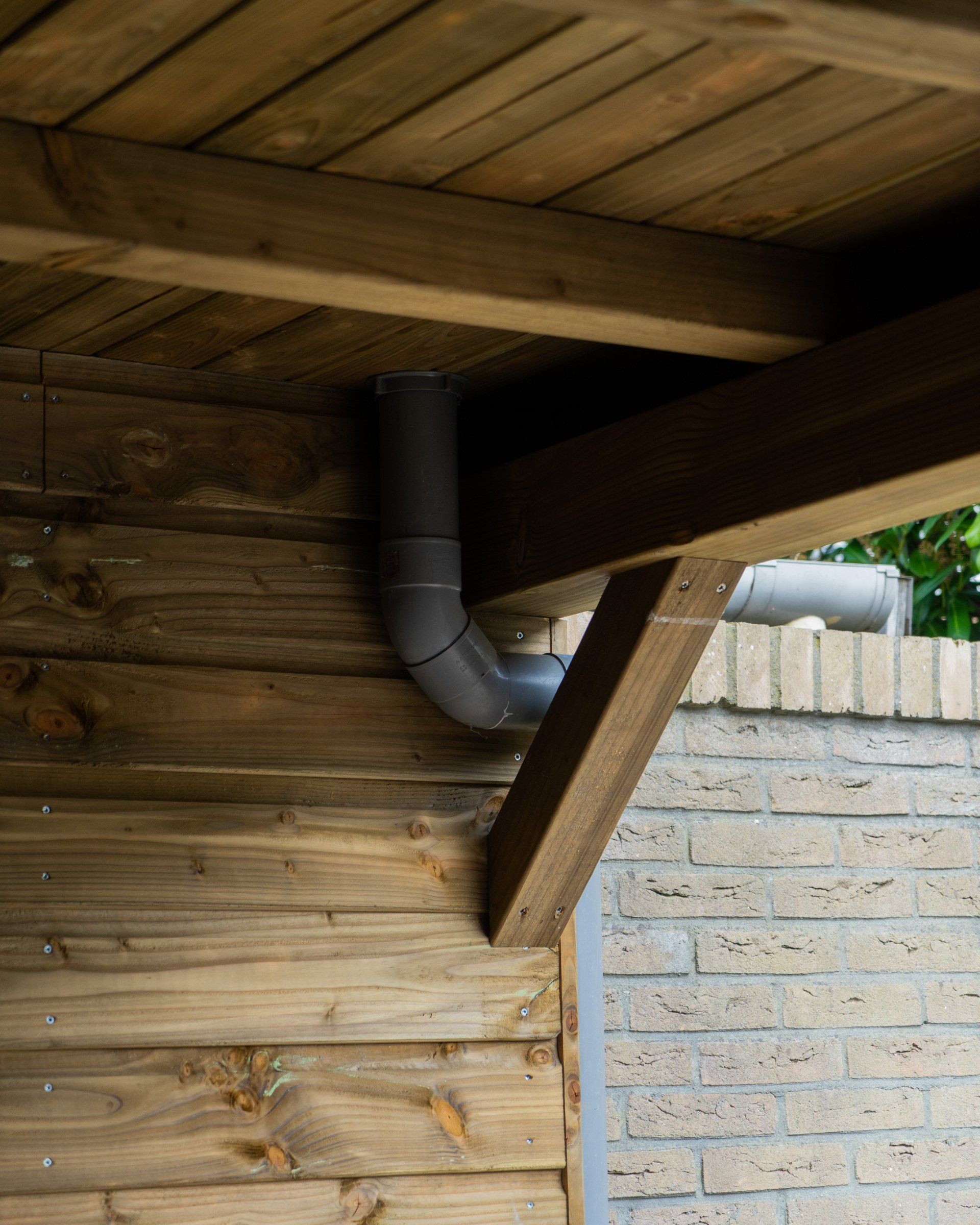 Grijze PVC-buis met bochten, gemonteerd op een houten constructie tegen een bakstenen muur.