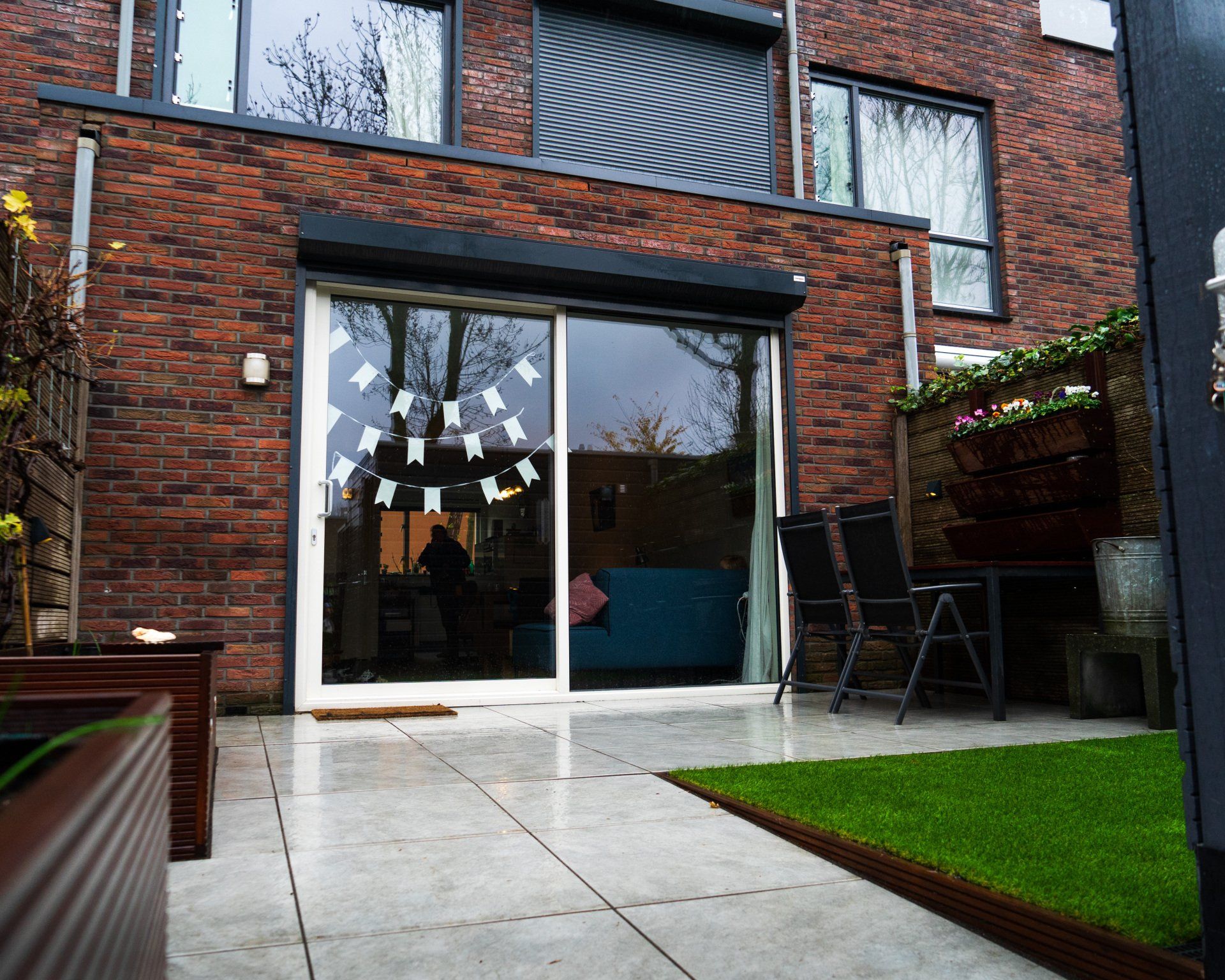 Achtertuin met bakstenen huis, schuifdeuren van glas, terras en een klein stukje groen gazon.