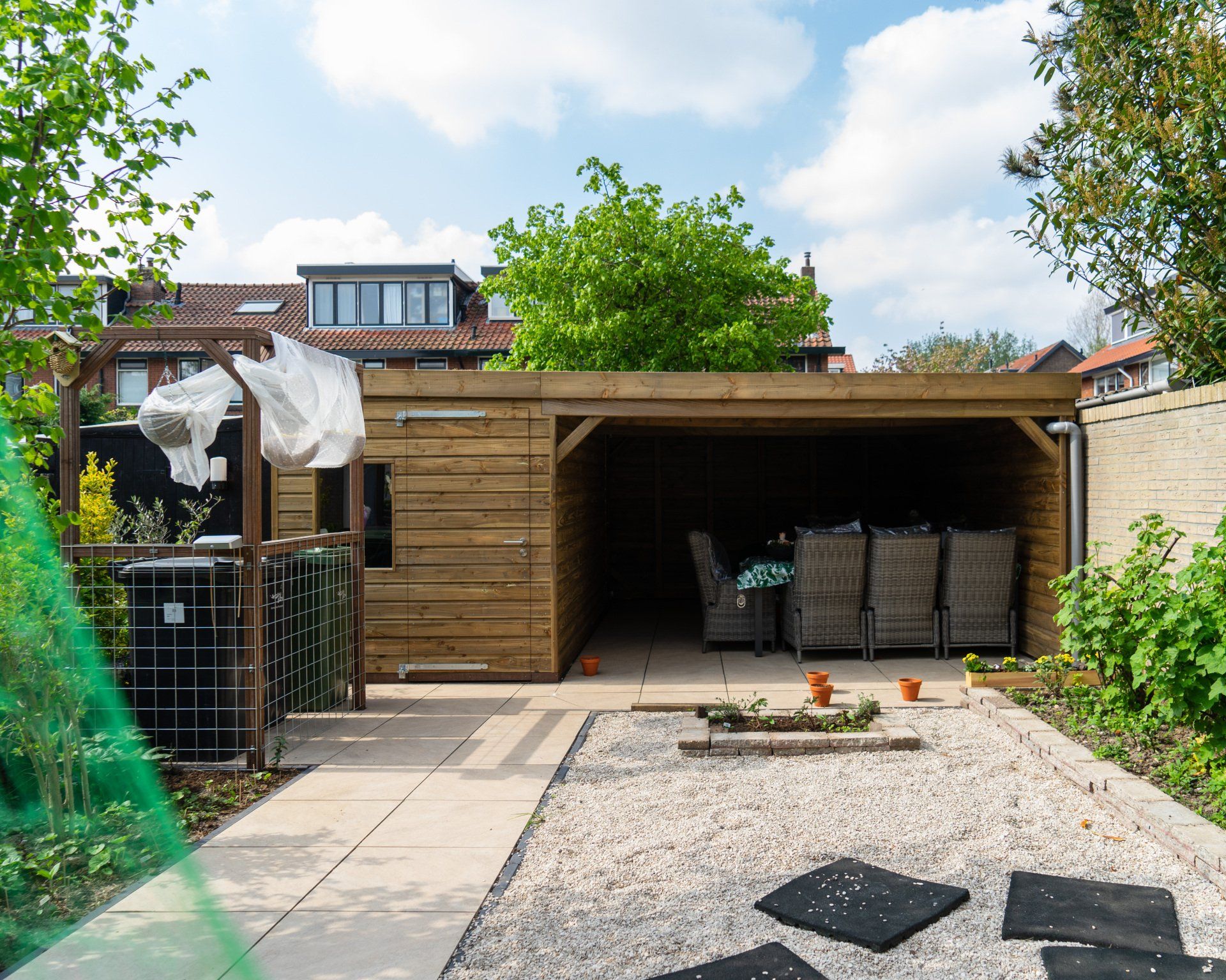 Houten schuur in een achtertuin met terrasmeubilair en een pad van grind en stenen.