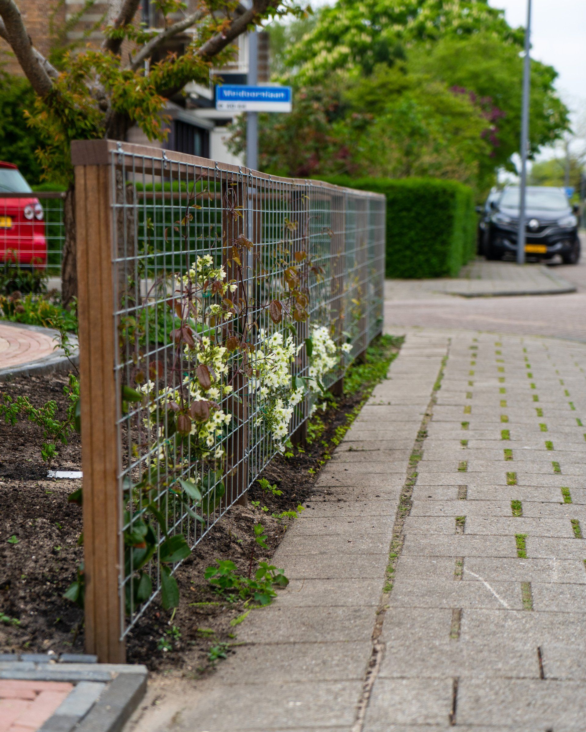 Een draadhek met bloemen langs een stoep naast een straat met auto's en een verkeersbord.