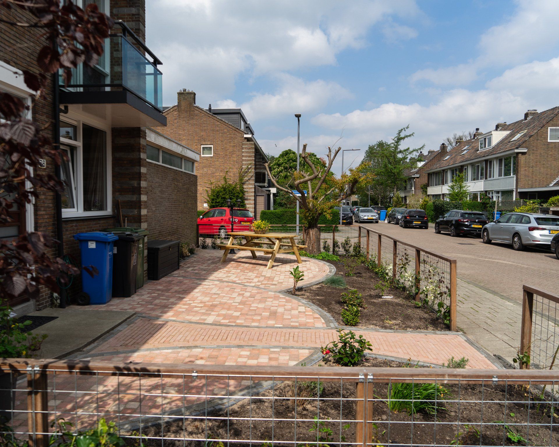 Straatbeeld met een bakstenen stoep, picknicktafel, beplanting en geparkeerde auto's op een zonnige dag.