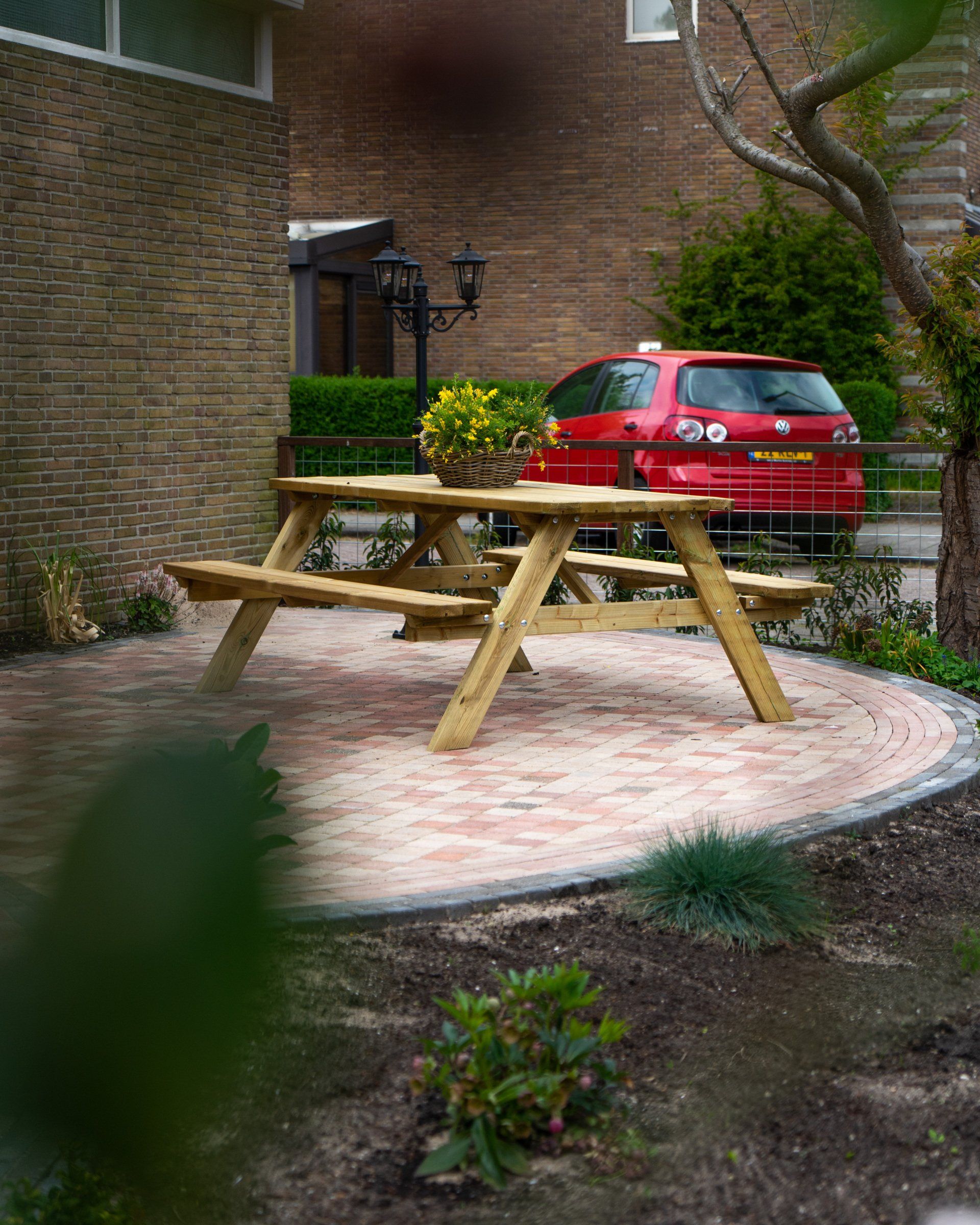 Een houten picknicktafel op een stenen terras, met een rode auto ernaast geparkeerd.