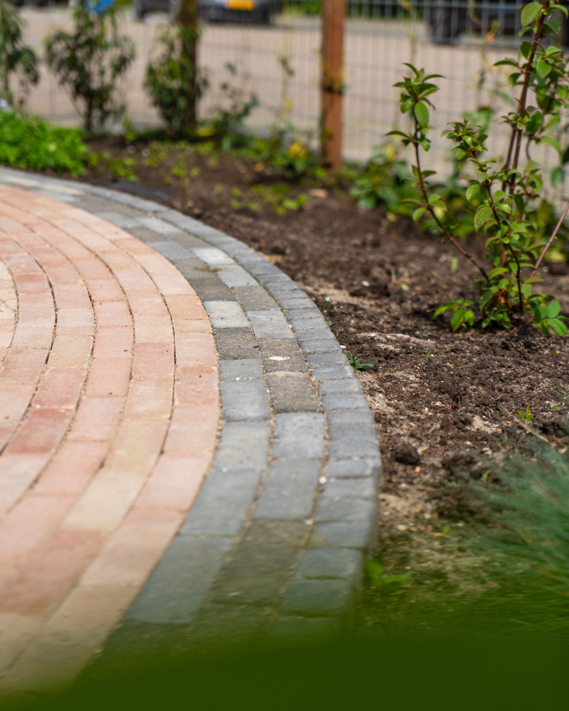 Een gebogen pad van rode en grijze bakstenen, omzoomd door aarde en planten.