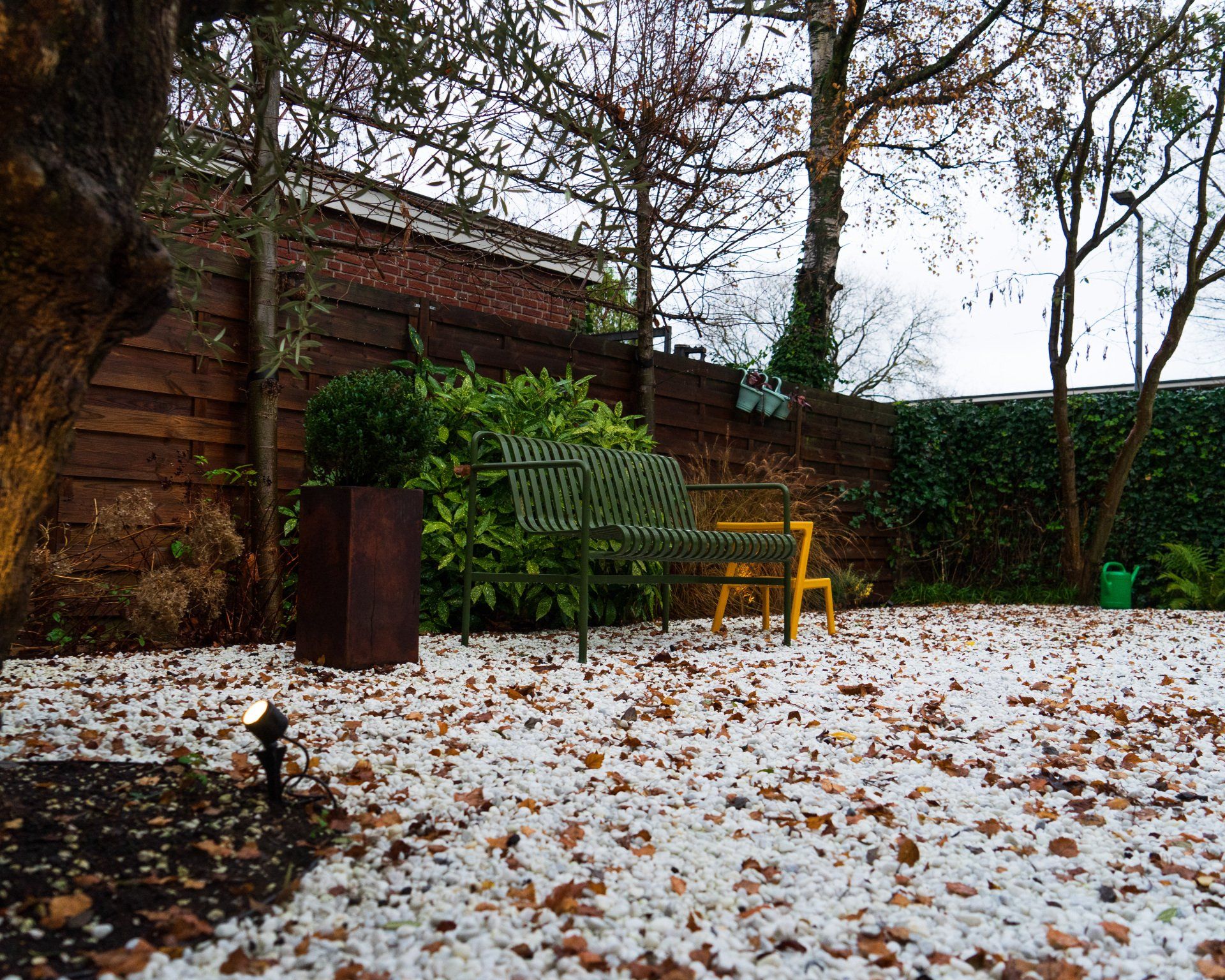 Tuin met wit grind, een bruin hek en een groen-gele bank.