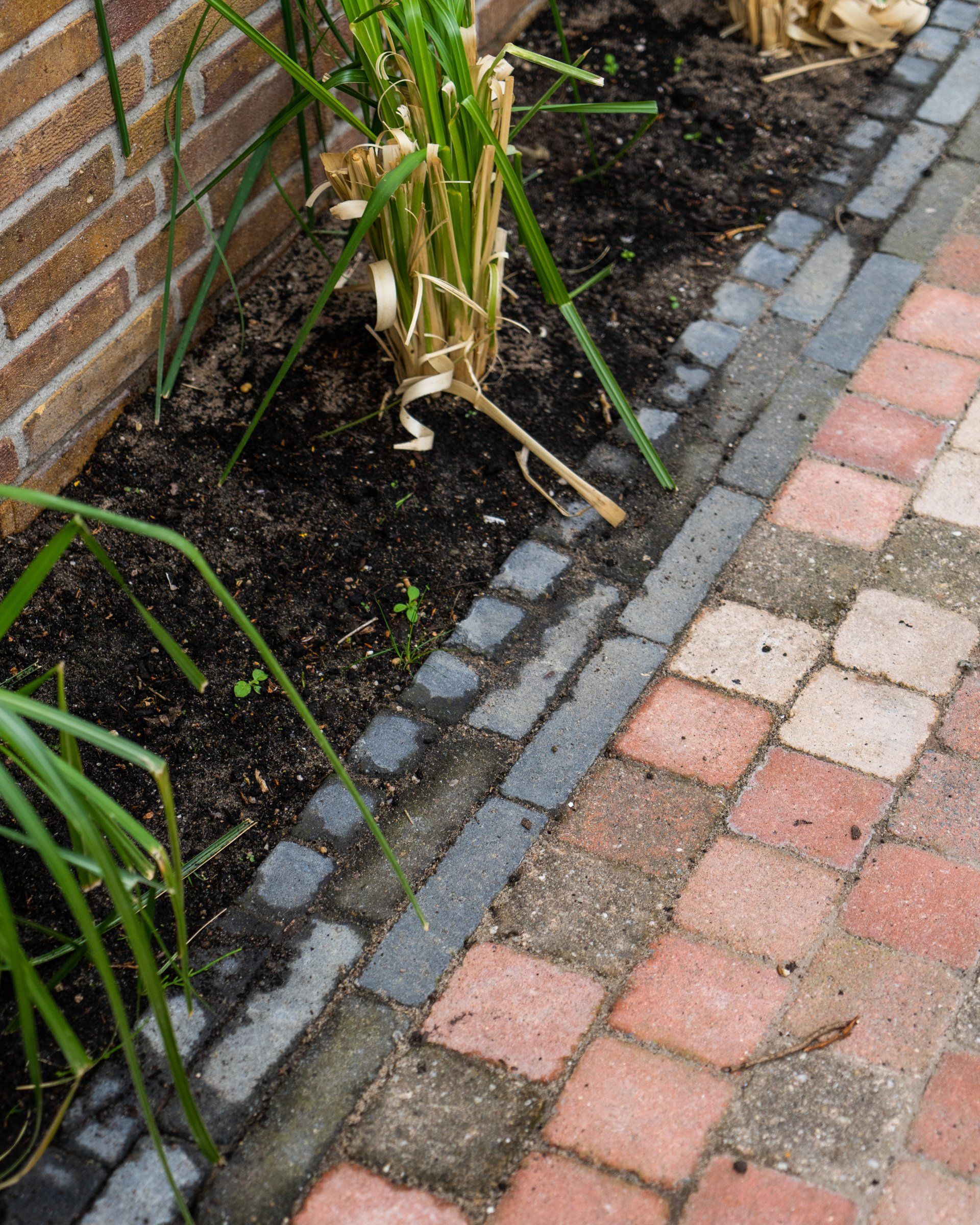 Een pad van bakstenen en een bloembed met donkere aarde, omzoomd door grijze bakstenen naast een rode bakstenen muur.