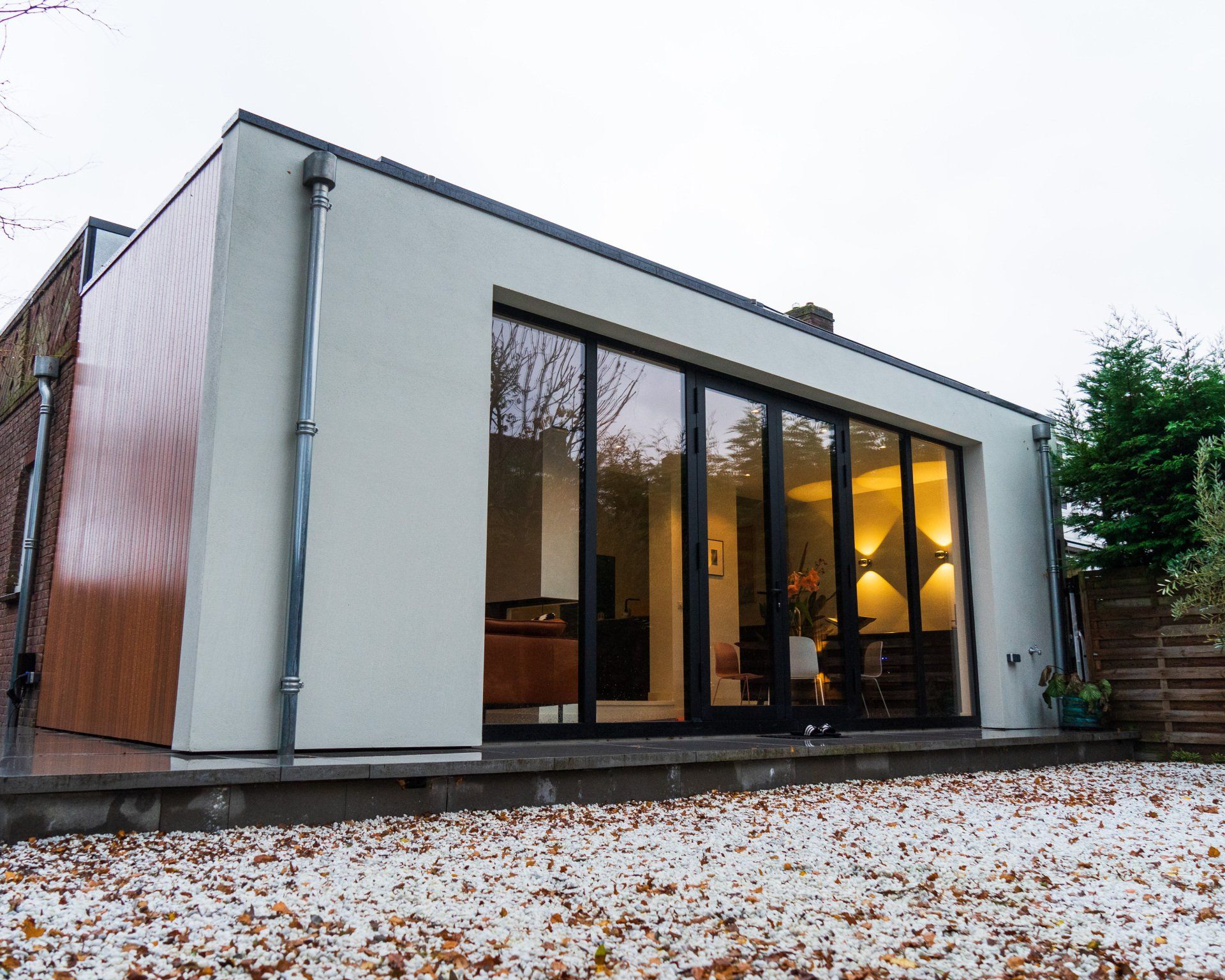 Moderne, witgekalkte aanbouw met grote glazen deuren. De zijkant is bekleed met houten panelen en staat op een grindpad met herfstbladeren.