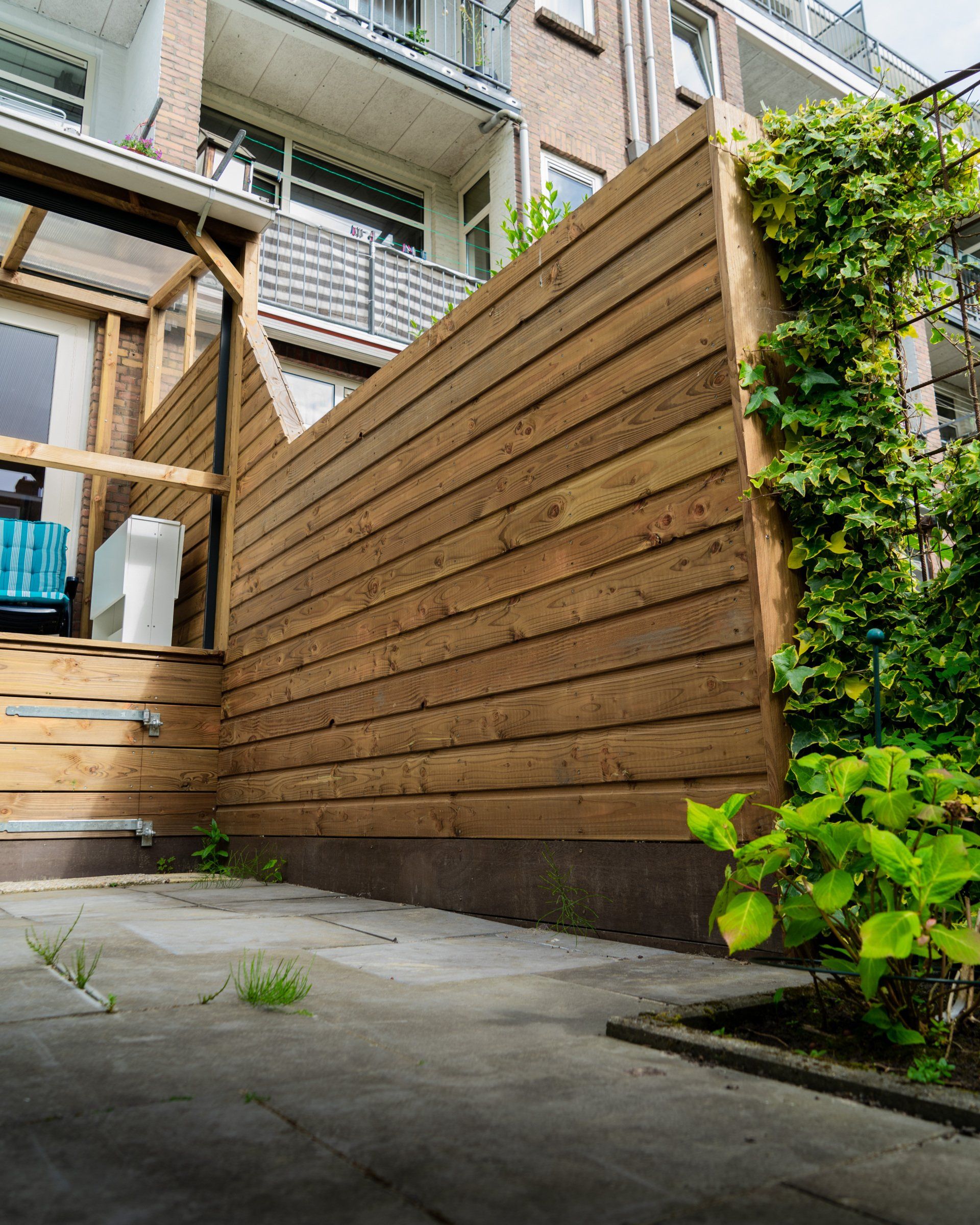 Een houten schutting staat langs een gebouw. Groene planten groeien ernaast.