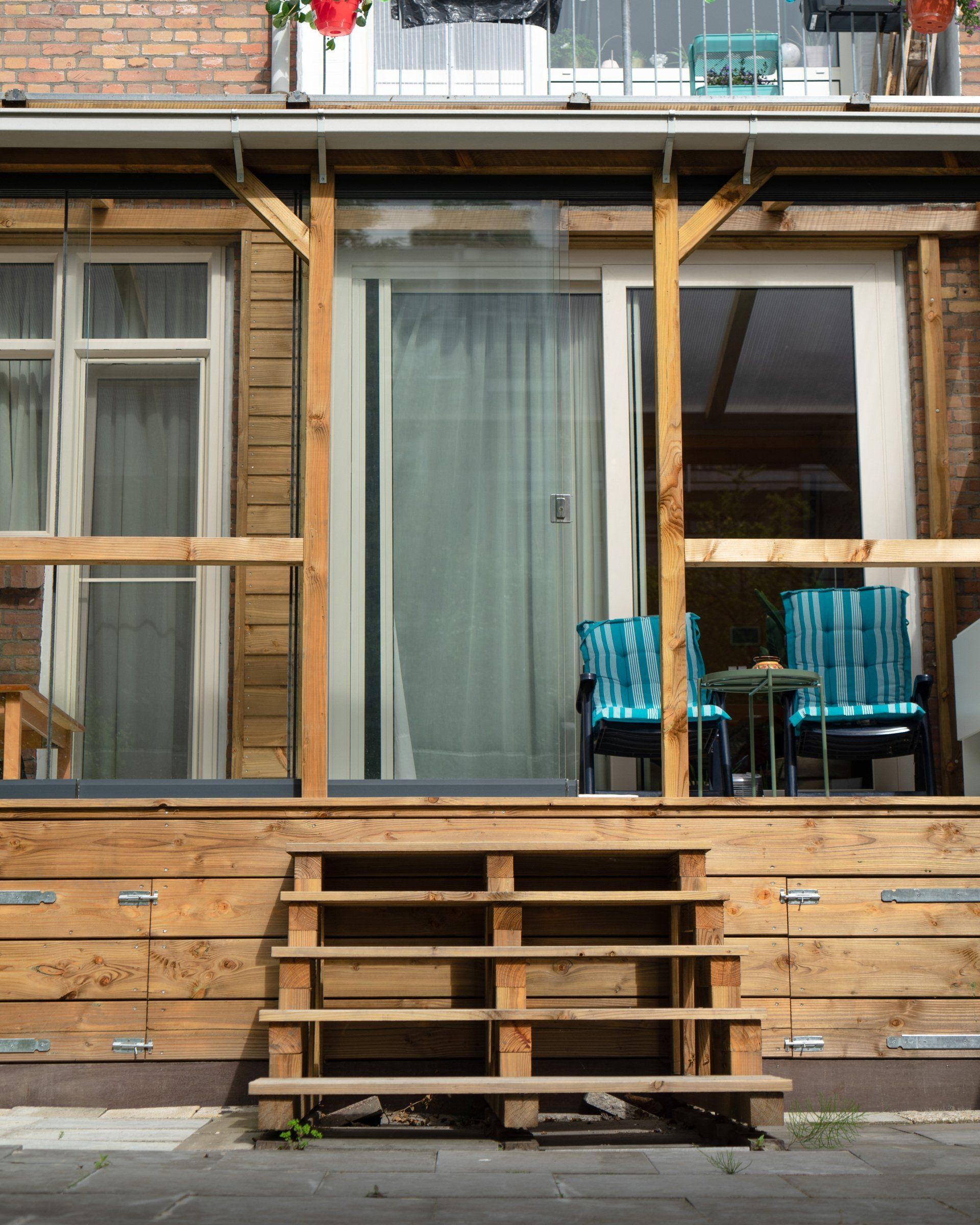Houten veranda met trap, blauwe stoelen, schuifdeur en bovenbalkon met rode lantaarns.