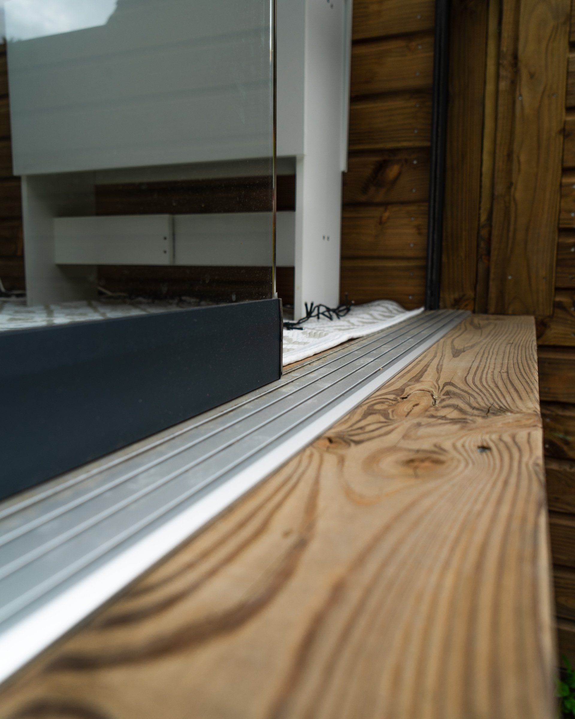 Houten terras met metalen rails naast een glazen raam. Bruine houten gevelbekleding op de achtergrond.