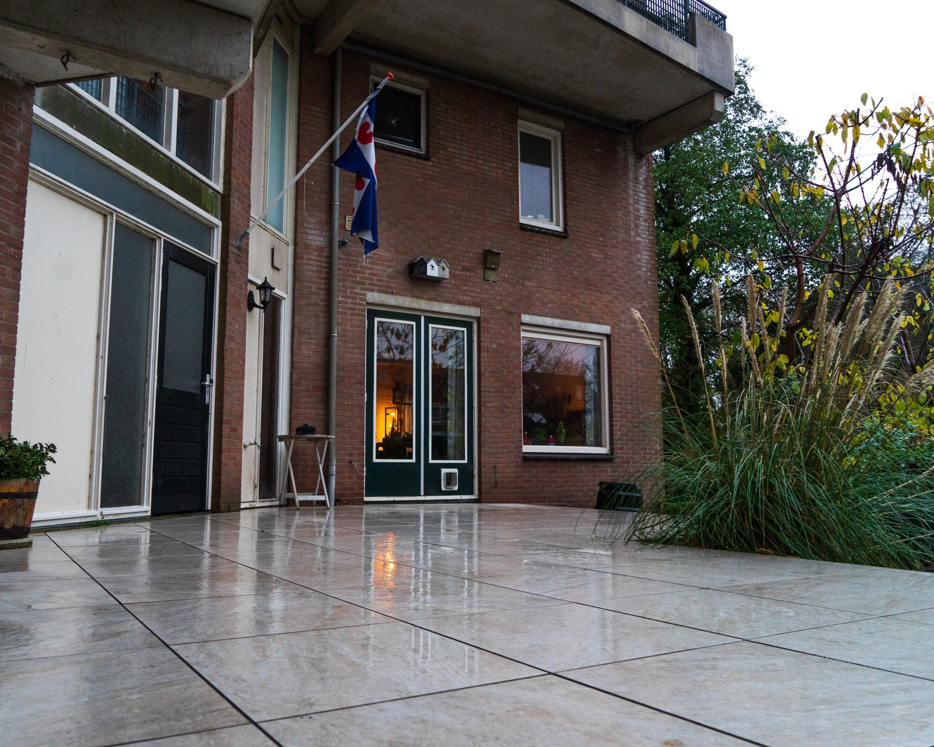 Bakstenen gebouw met Nederlandse vlag, glazen deuren die het binnenlicht reflecteren op de natte patio.