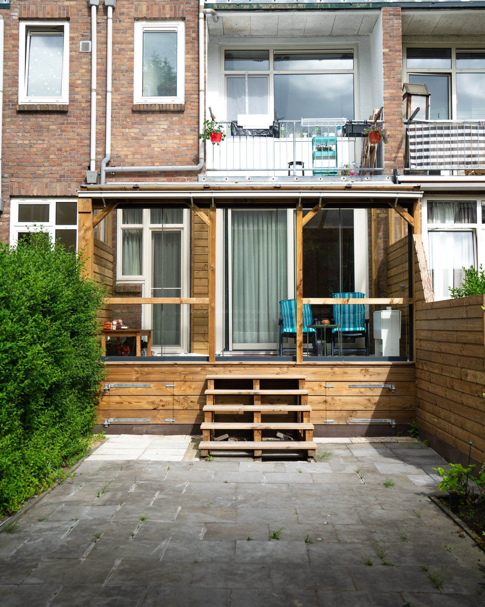 Houten terras met een trap die naar een glazen schuifdeur leidt. Op de achtergrond een bakstenen gebouw.