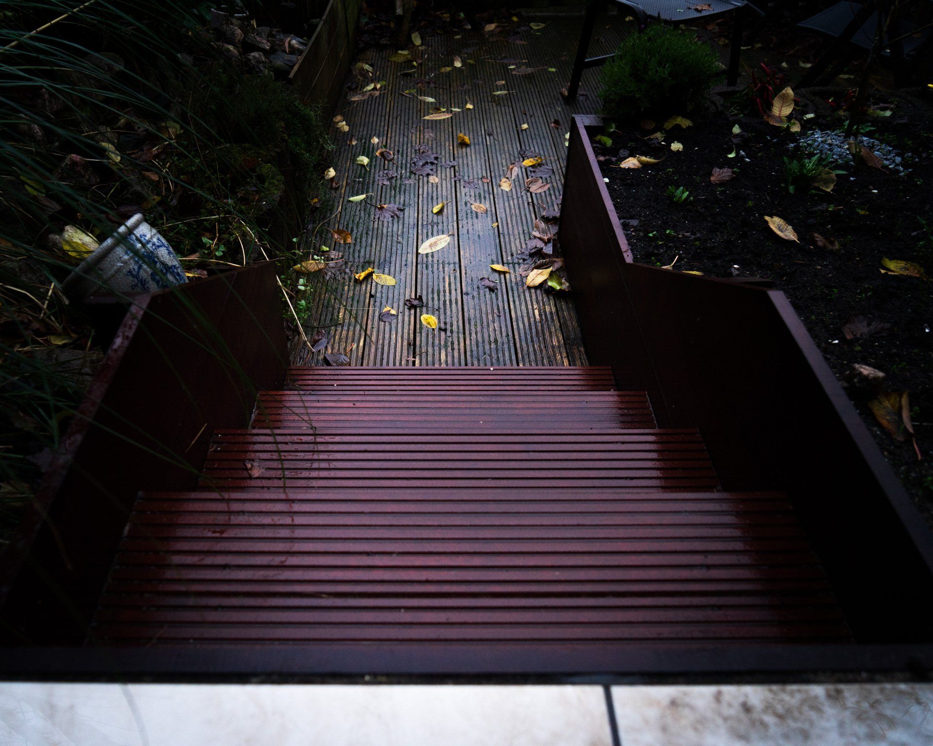 Houten trappen en een pad leiden naar beneden, naar een tuin die nat is van de regen en gevallen bladeren.