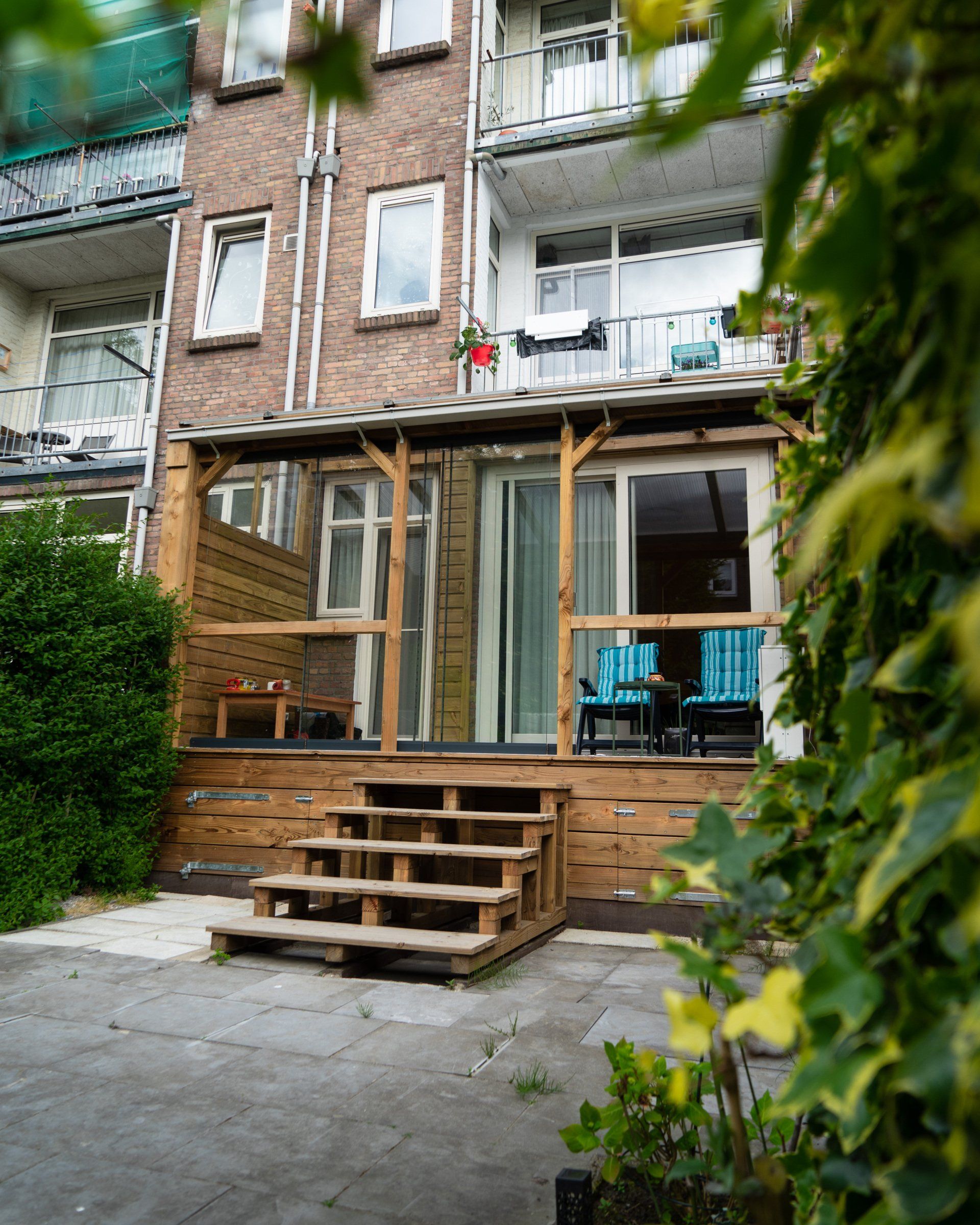 Houten terras met trap, grenzend aan een bakstenen gebouw. Het terras heeft een dak en een glazen schuifdeur.