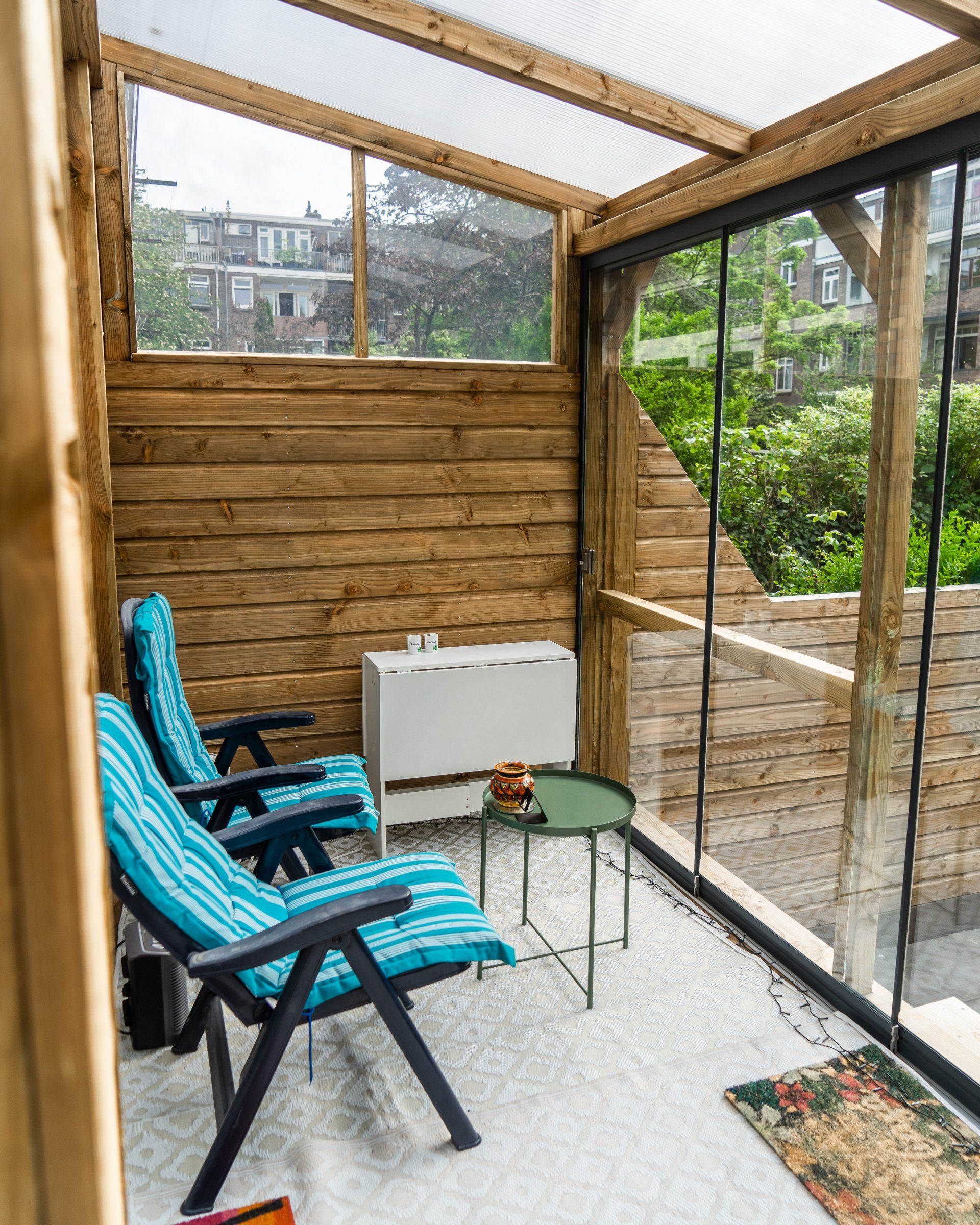 Een buitenterras met houten wanden, twee blauwe loungestoelen, glazen panelen, een tafeltje en een klein wit kastje.