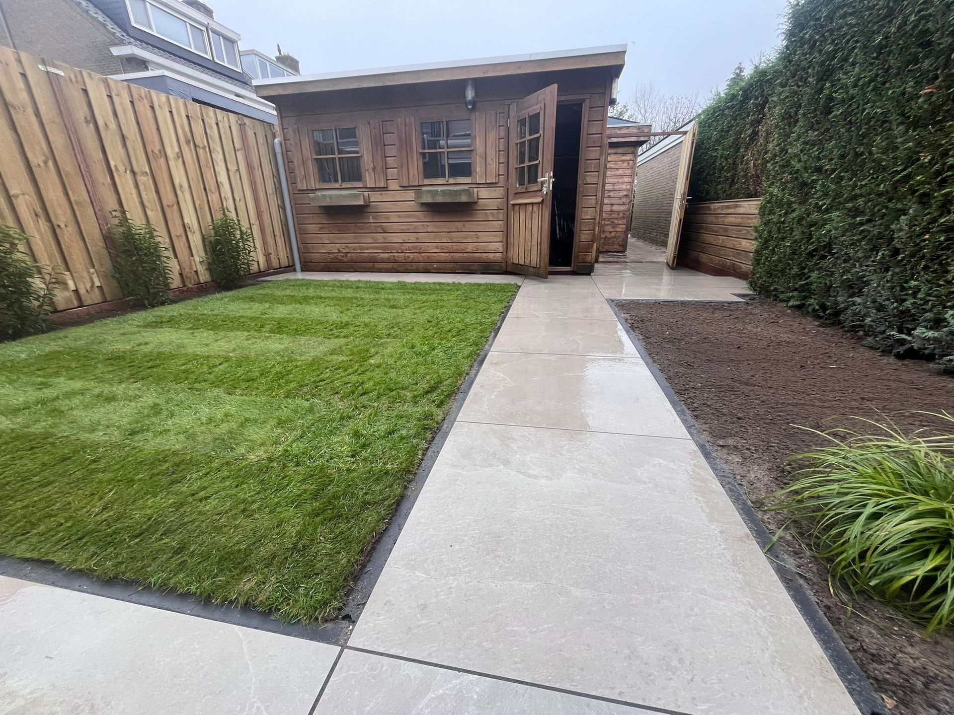 Kleine achtertuin met een houten schuur, groen gazon, bestraat pad en hagen.