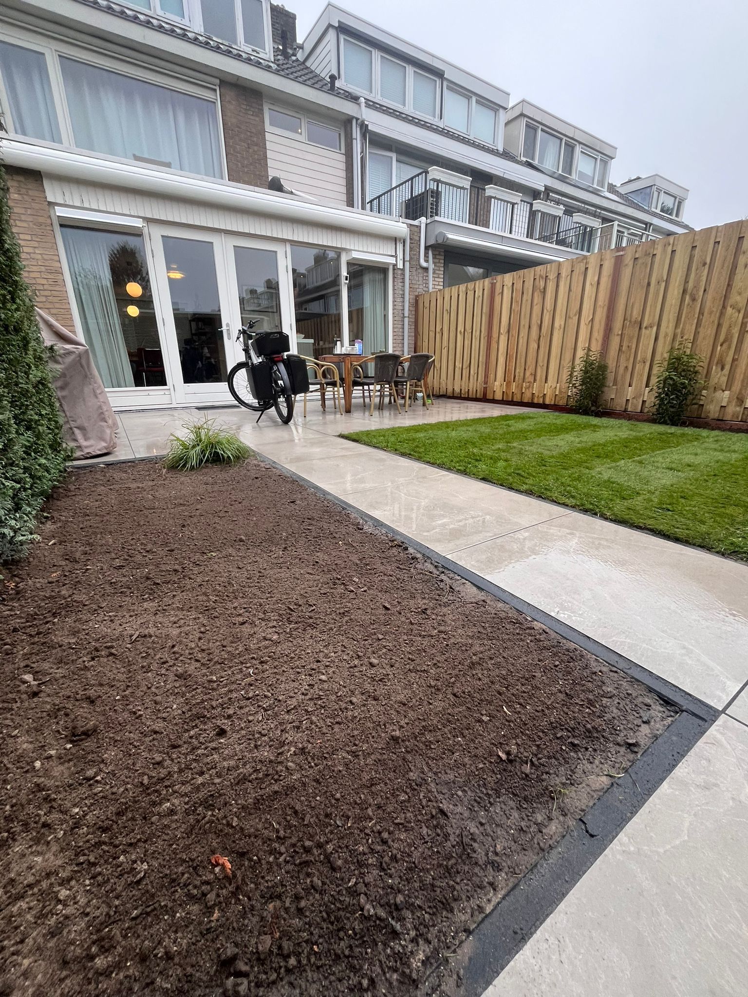 Een achtertuin met een bloembed, gazon, betonnen pad, houten schutting en een rij rijtjeshuizen.