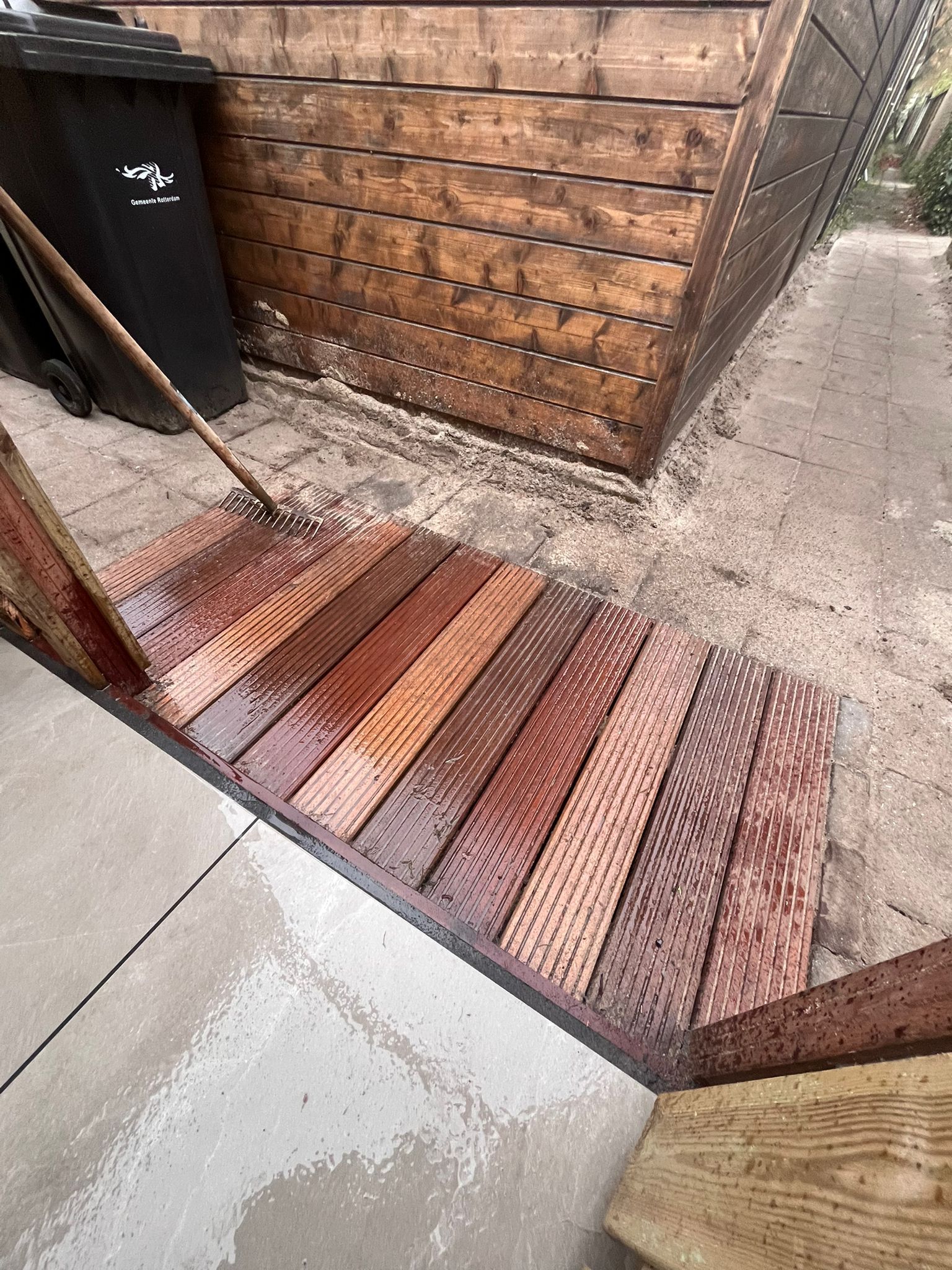 Houten hellingbaan met natte, veelkleurige planken die naar een bruine houten constructie leidt. De hellingbaan is afgewerkt met beton.