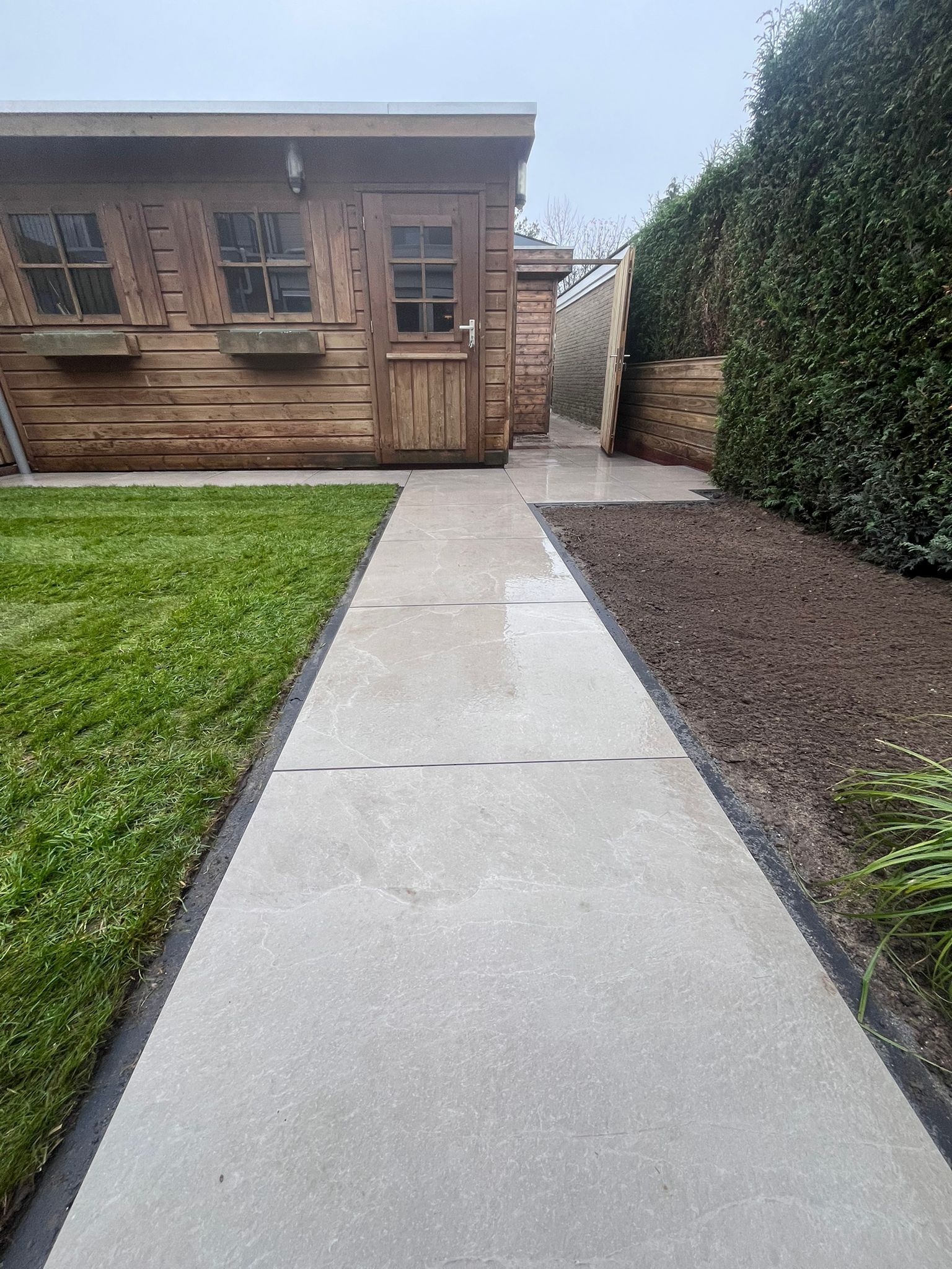 Een betonnen pad leidt naar een houten schuur, geflankeerd door gras aan de ene kant en een bloembed aan de andere kant.