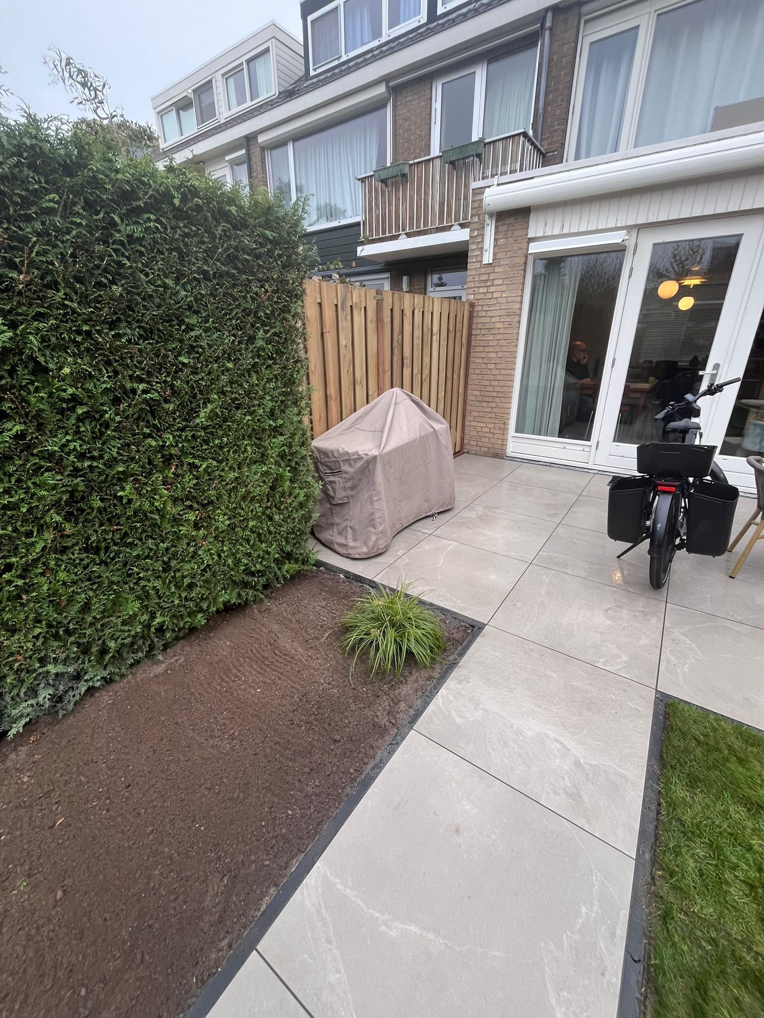 Terras met een overdekte barbecue, omgeven door een heg, een schutting en een gebouw met glazen deuren. Een fiets staat in de buurt geparkeerd.