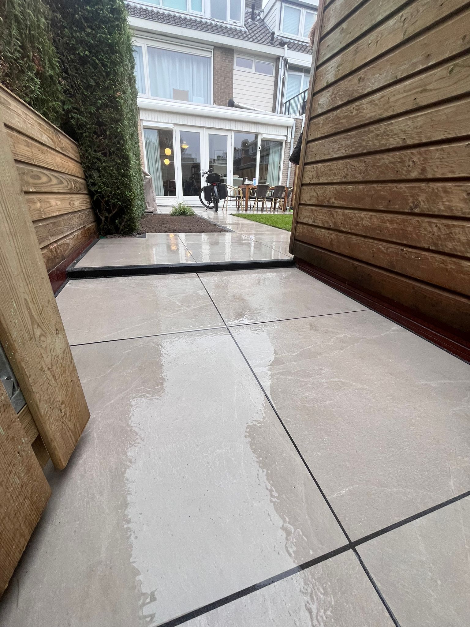 Een nat, lichtgekleurd terras met donkere lijnen, begrensd door houten muren. Het uitzicht leidt naar een gebouw met glazen deuren.