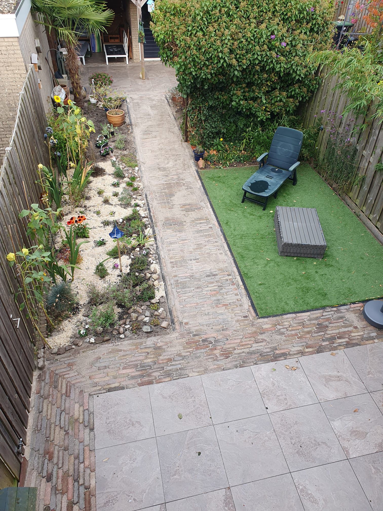 Bovenaanzicht van een kleine tuin met een pad, bloemen, een tuinstoel en bakstenen muren.