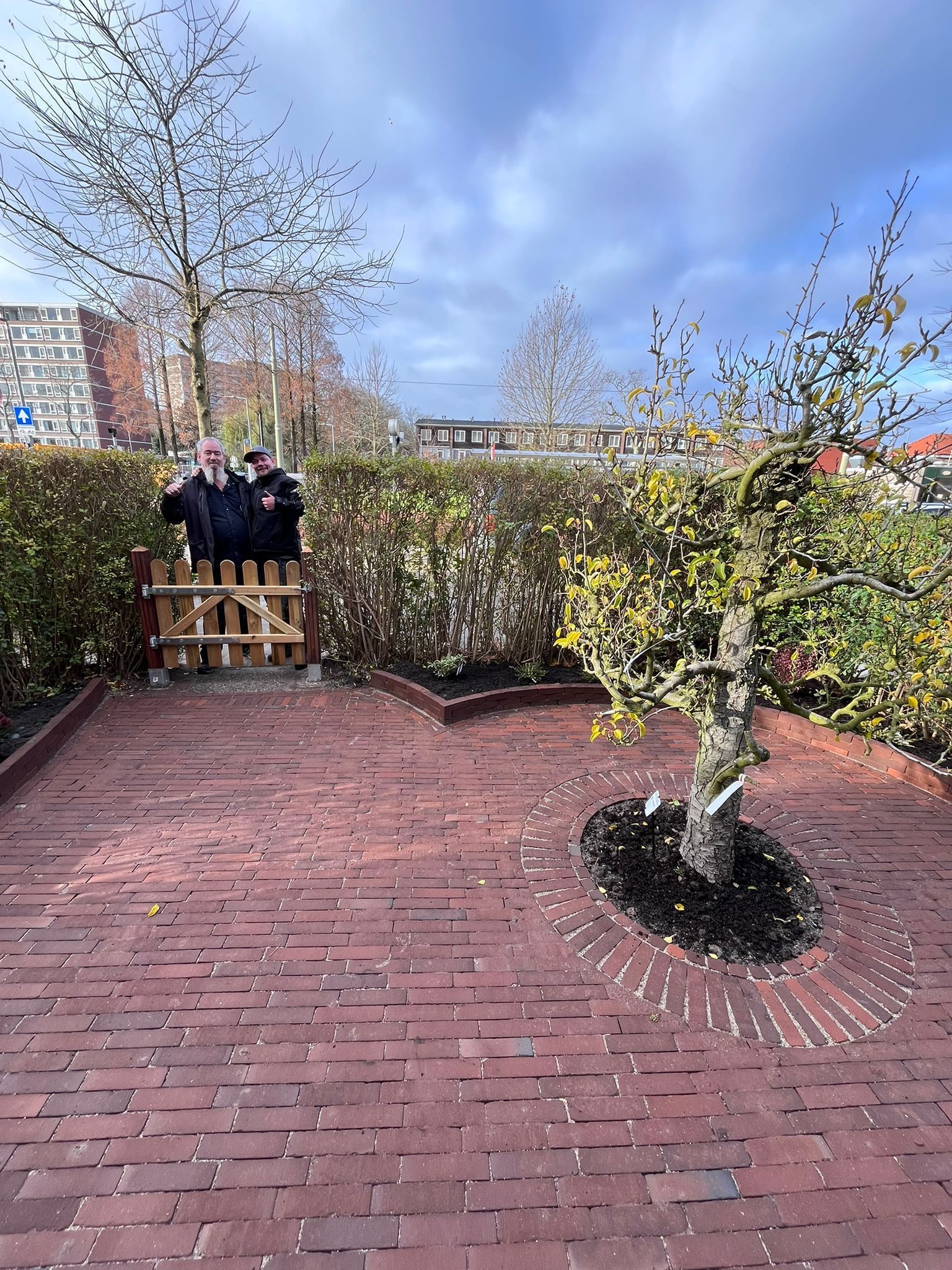 Een bakstenen pad leidt naar een kleine tuin met een jonge boom, een haag en twee figuren bij een poort.