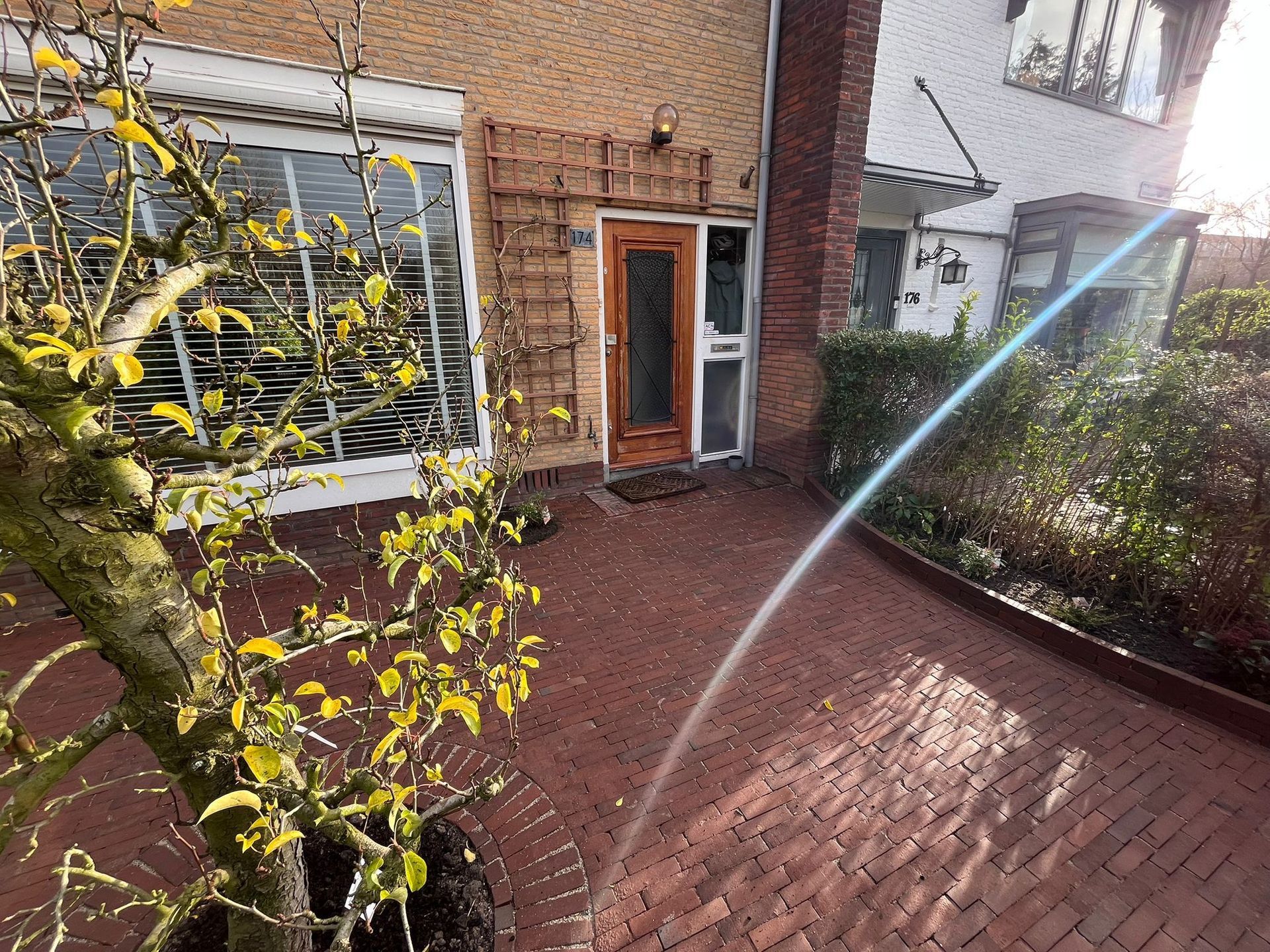 De ingang van een bakstenen huis met een kleine boom op de voorgrond. De deur is zichtbaar. Zonlicht schijnt over de bakstenen oprit.