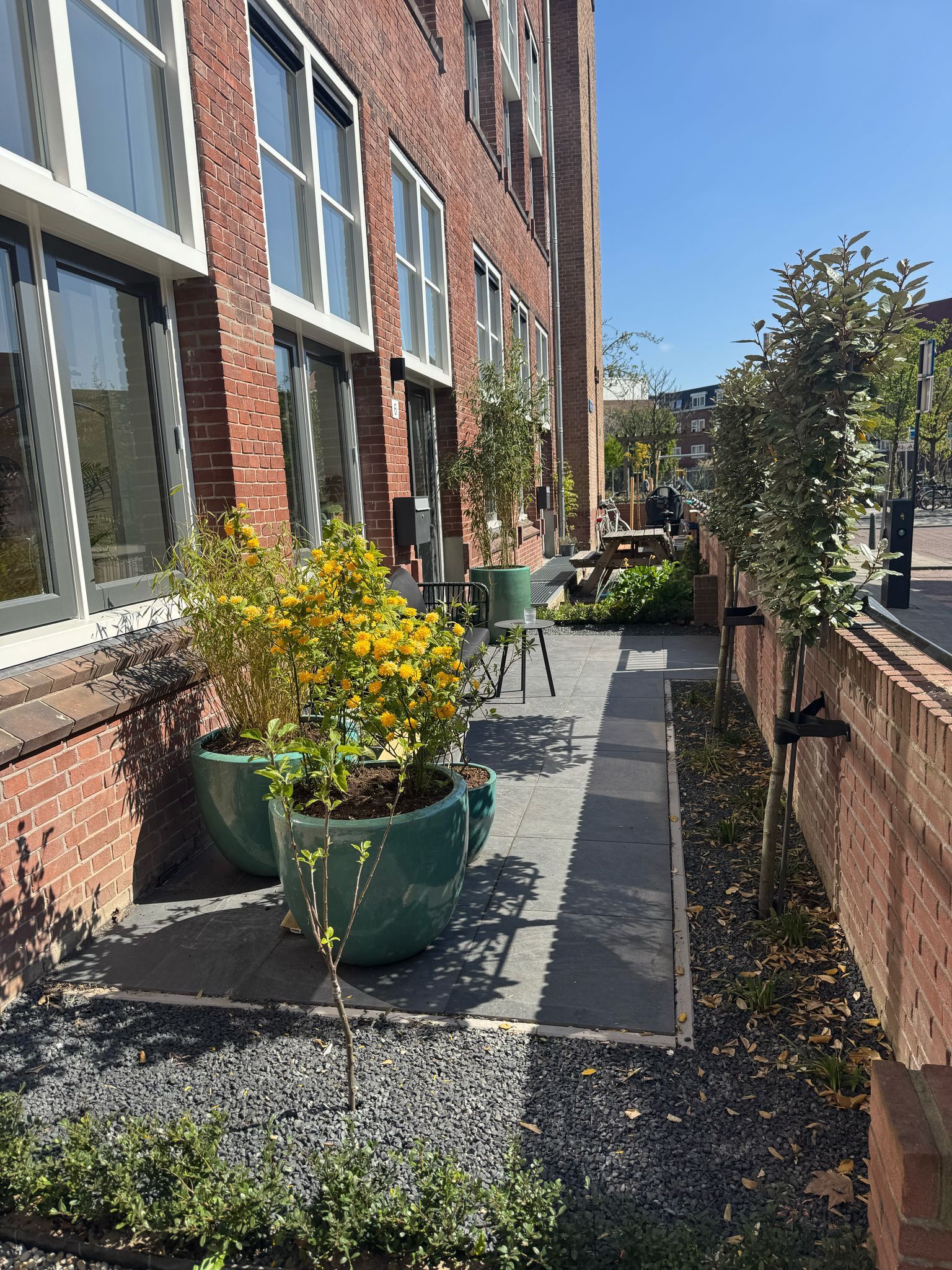 Een bakstenen gebouw met een smal, schaduwrijk terras met potplanten en een kleine zithoek.