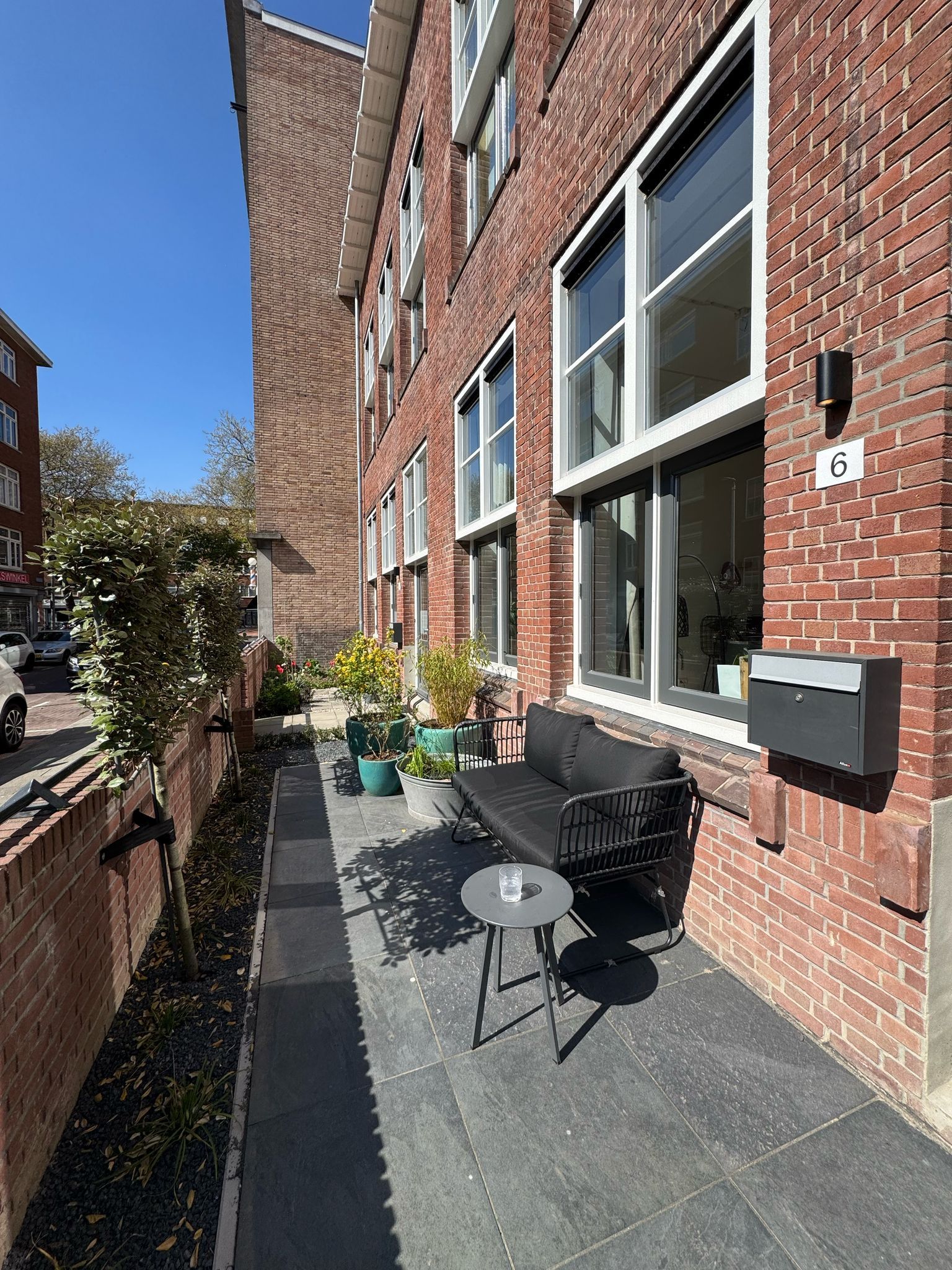 Bakstenen gebouw met een zithoek, een kleine tafel en potplanten. Het gebouw heeft meerdere ramen.